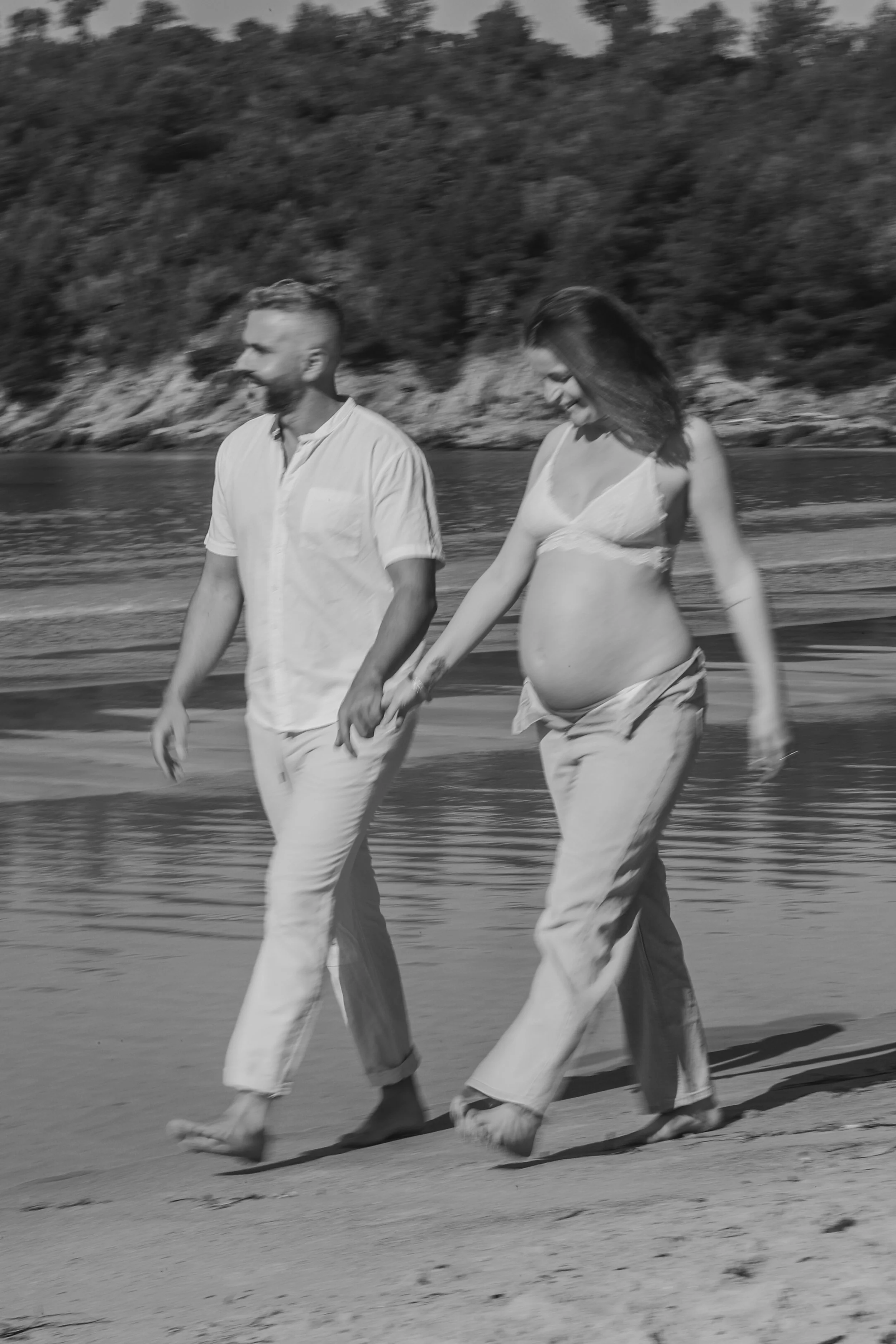 Un couple marchant main dans la main sur la plage, la femme enceinte porte un haut clair et un pantalon, l'homme porte une chemise et un pantalon, tous deux pieds nus, avec une mer et des collines en arrière-plan.