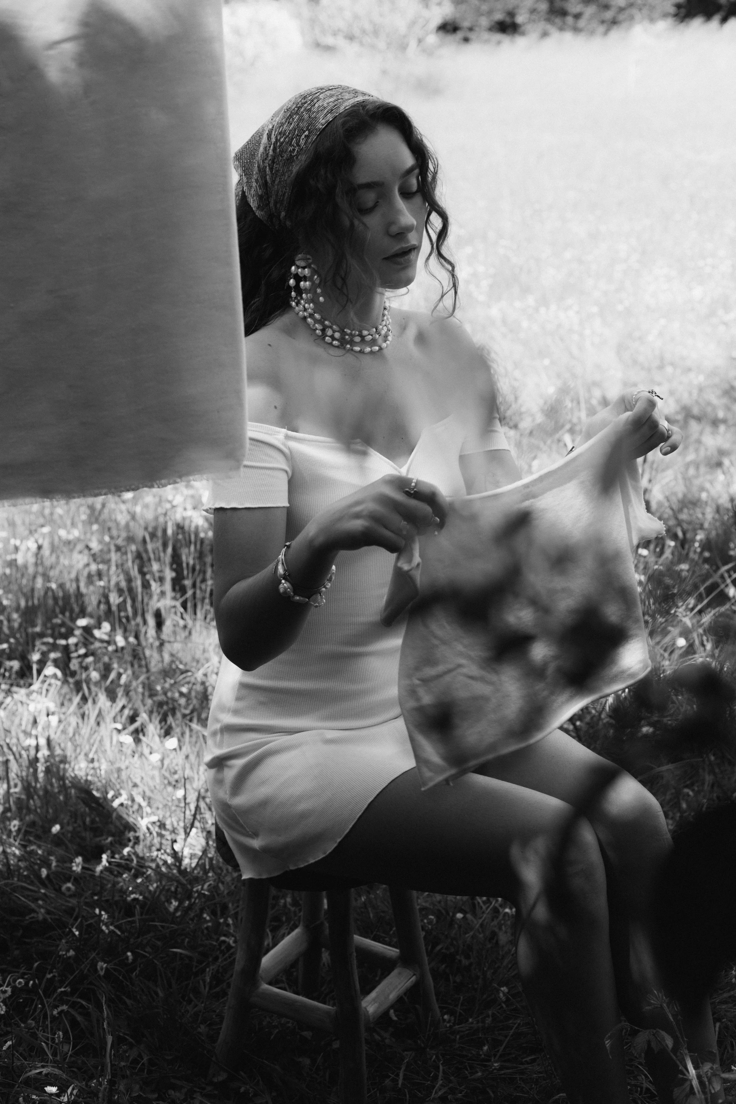 Une femme assise sur un tabouret en bois, portant une robe blanche, des bijoux et un foulard, dans un champ, regardant un chiffon qu'elle tient.