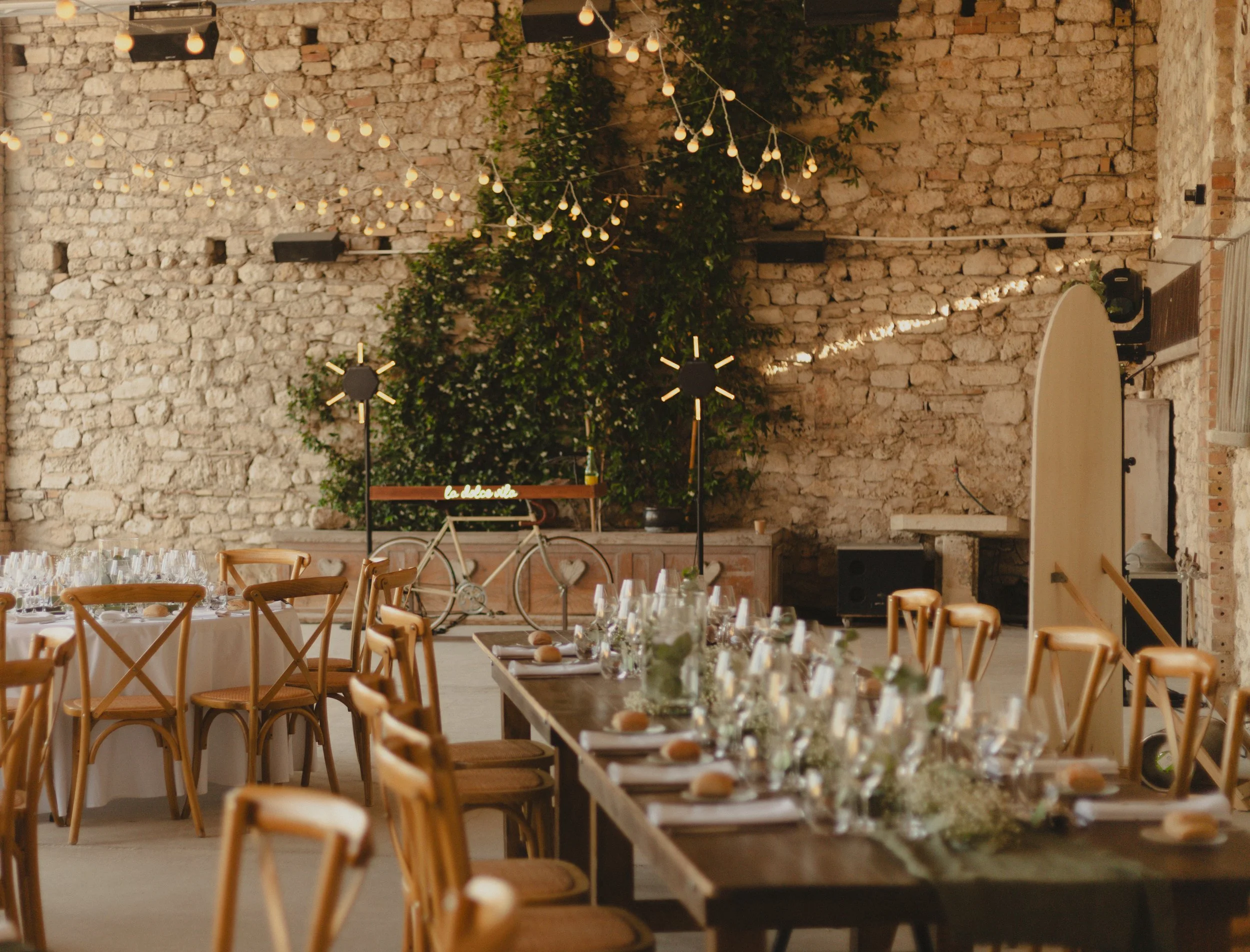 Une salle décorée pour un événement, avec tables garnies de verres et assiettes, chaises en bois, guirlandes lumineuses, un mur en pierre et un vélo décoratif avec une plaque "la dolce vita".
