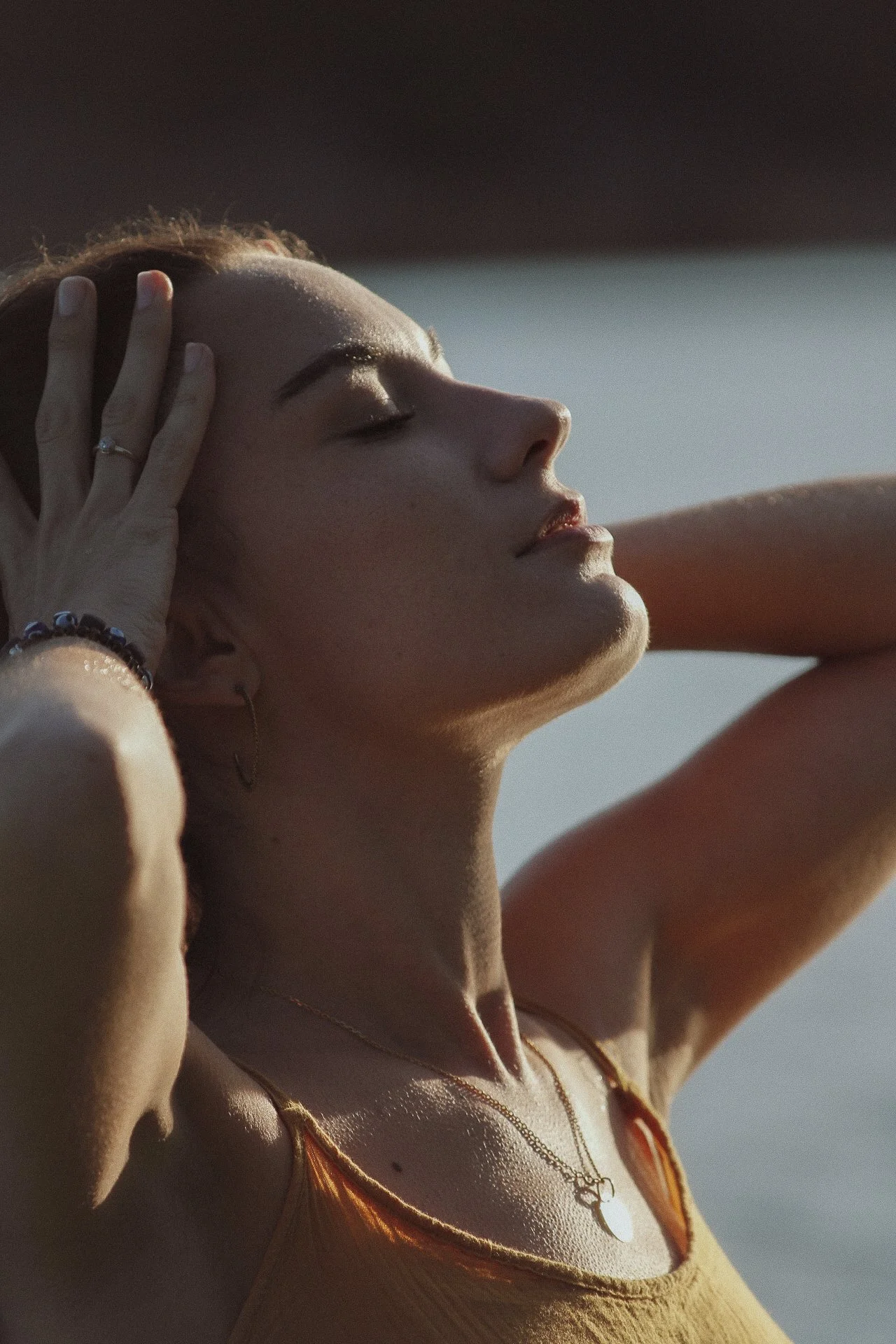 Jeune femme en détente avec yeux fermés, portant une chaîne avec un pendentif en forme de cœur, un earring, et un bracelet, en extérieur ensoleillé.