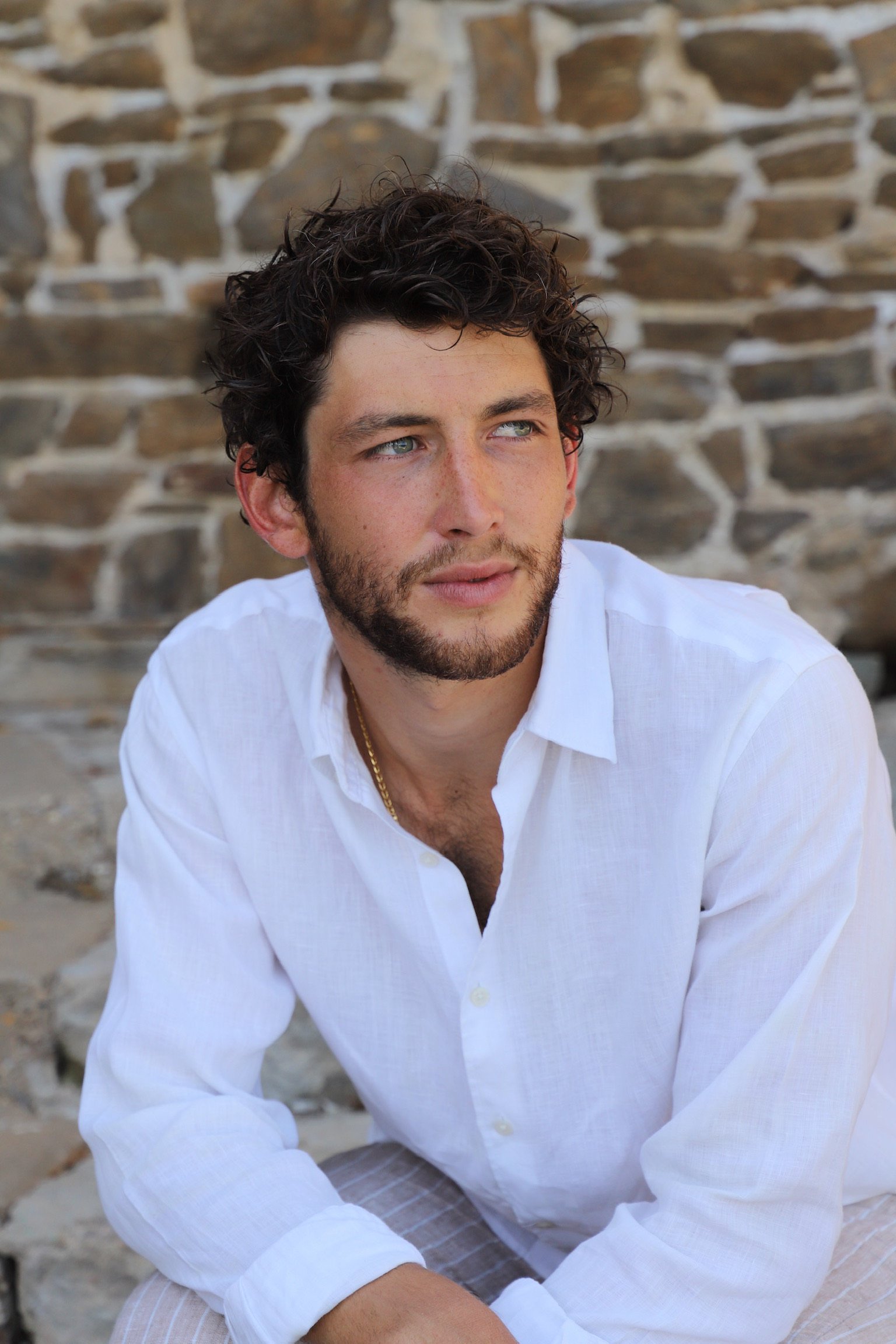 Jeune homme aux cheveux bouclés et à la barbe, portant une chemise blanche, assis devant un mur en pierre, regardant vers la droite.