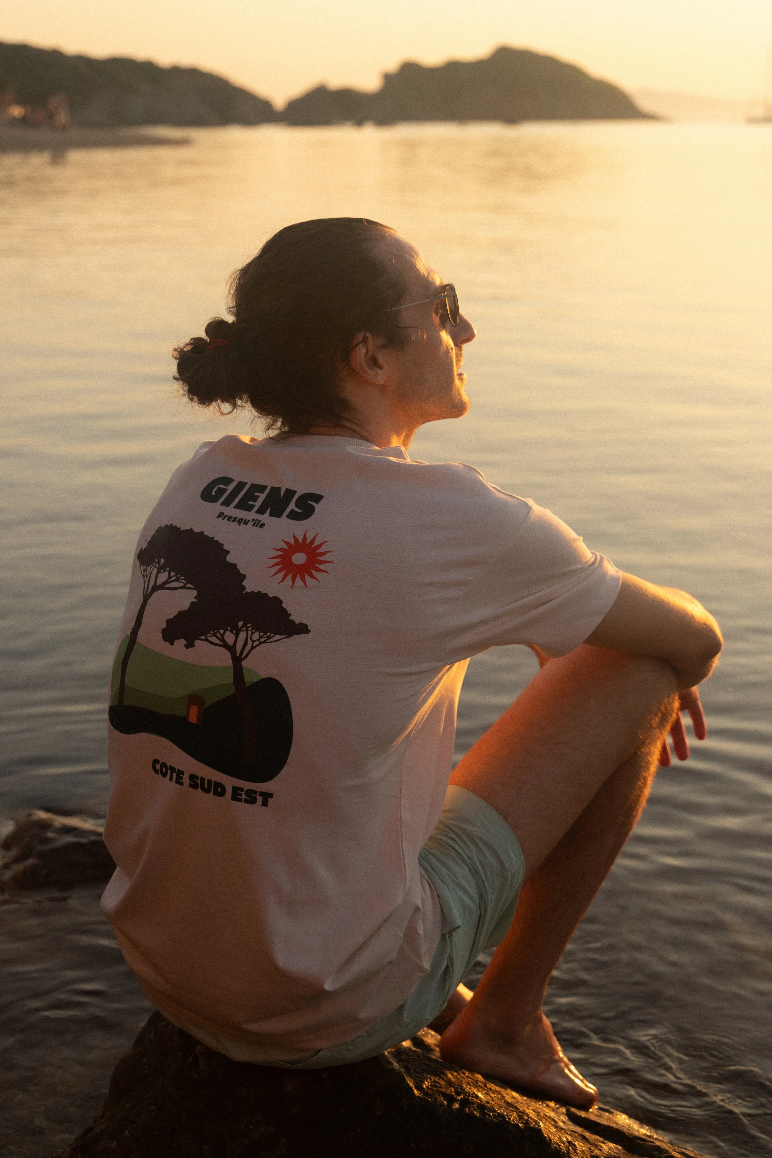 Un homme assis sur une pierre près de l'eau lors d'un coucher de soleil, portant un t-shirt blanc avec un design de deux arbres, un soleil rouge, et des textes en français, regardant vers l'horizon.