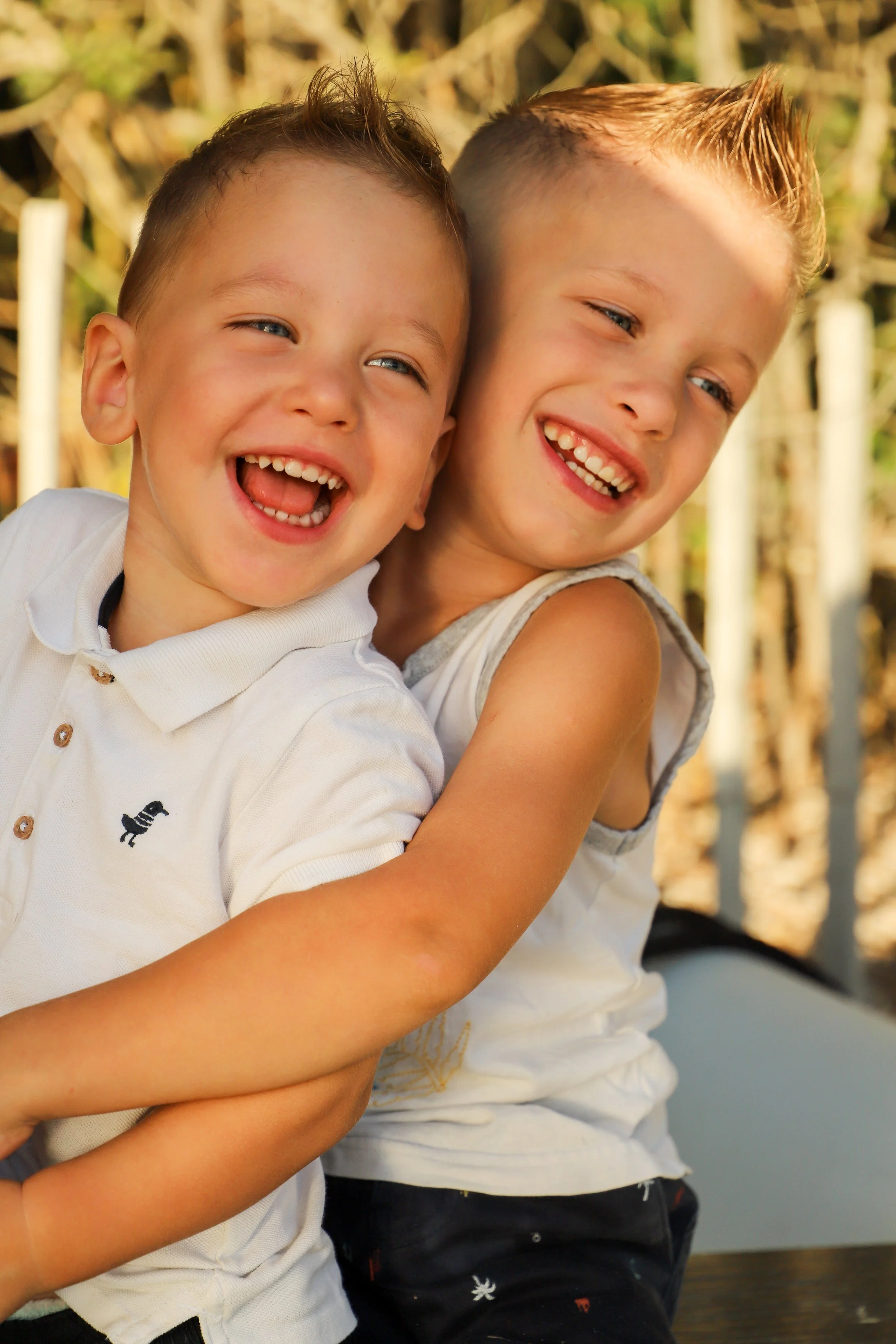 Deux jeunes garçons souriants, l'un avec une chemise blanche, l'autre avec un t-shirt sans manches, se font un câlin en plein air, au soleil, avec un fond flou de végétation.