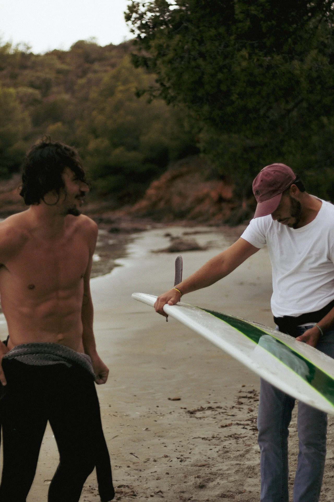 Deux jeunes hommes se tiennent sur une plage en discussion, l'un est torse nu, l'autre porte une casquette rose et tient une planche de surf.