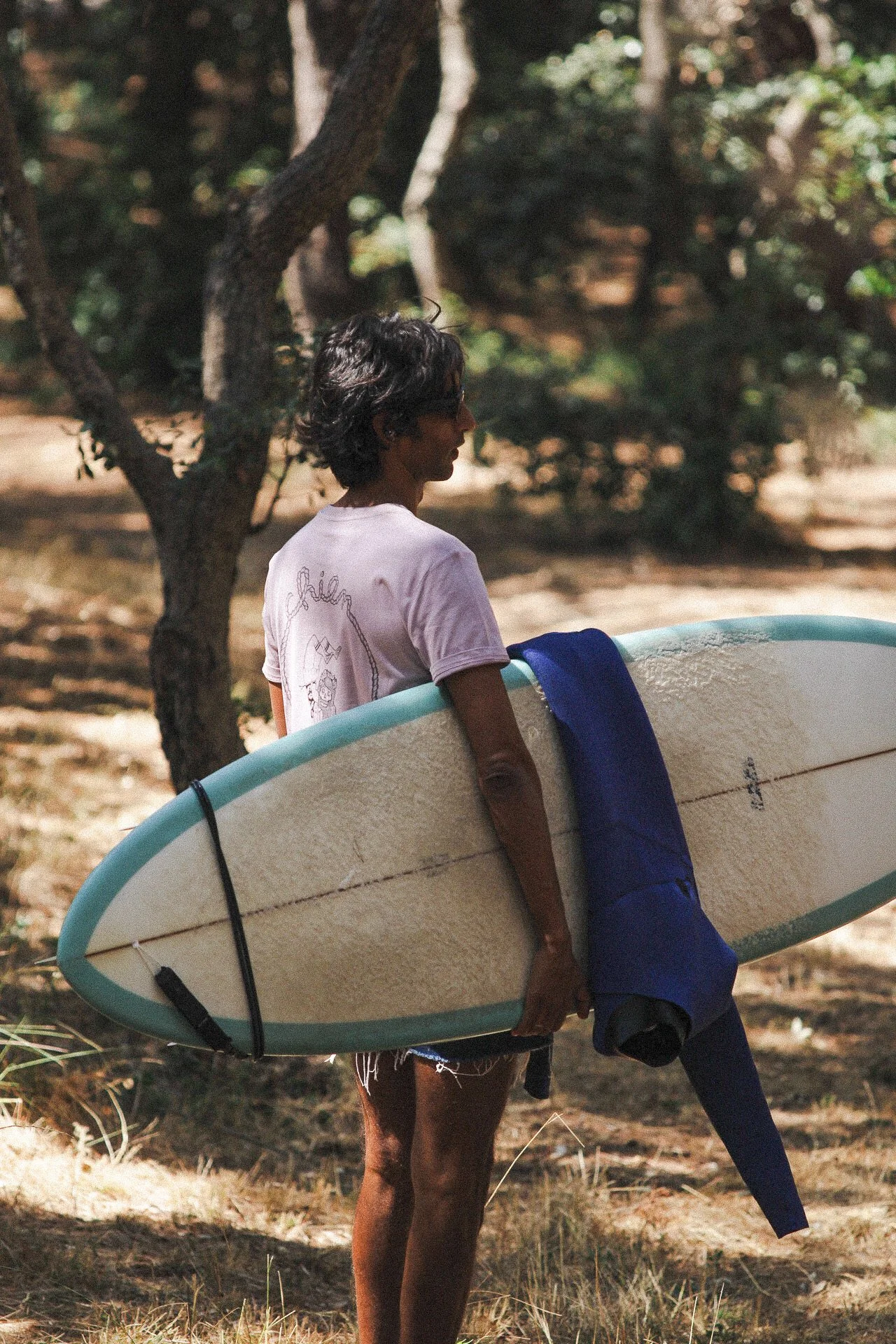 Femme debout dans un parc avec un skimboard enroulé sous le bras, portant une t-shirt blanche et des vêtements décontractés, entourée d'arbres et de nature.