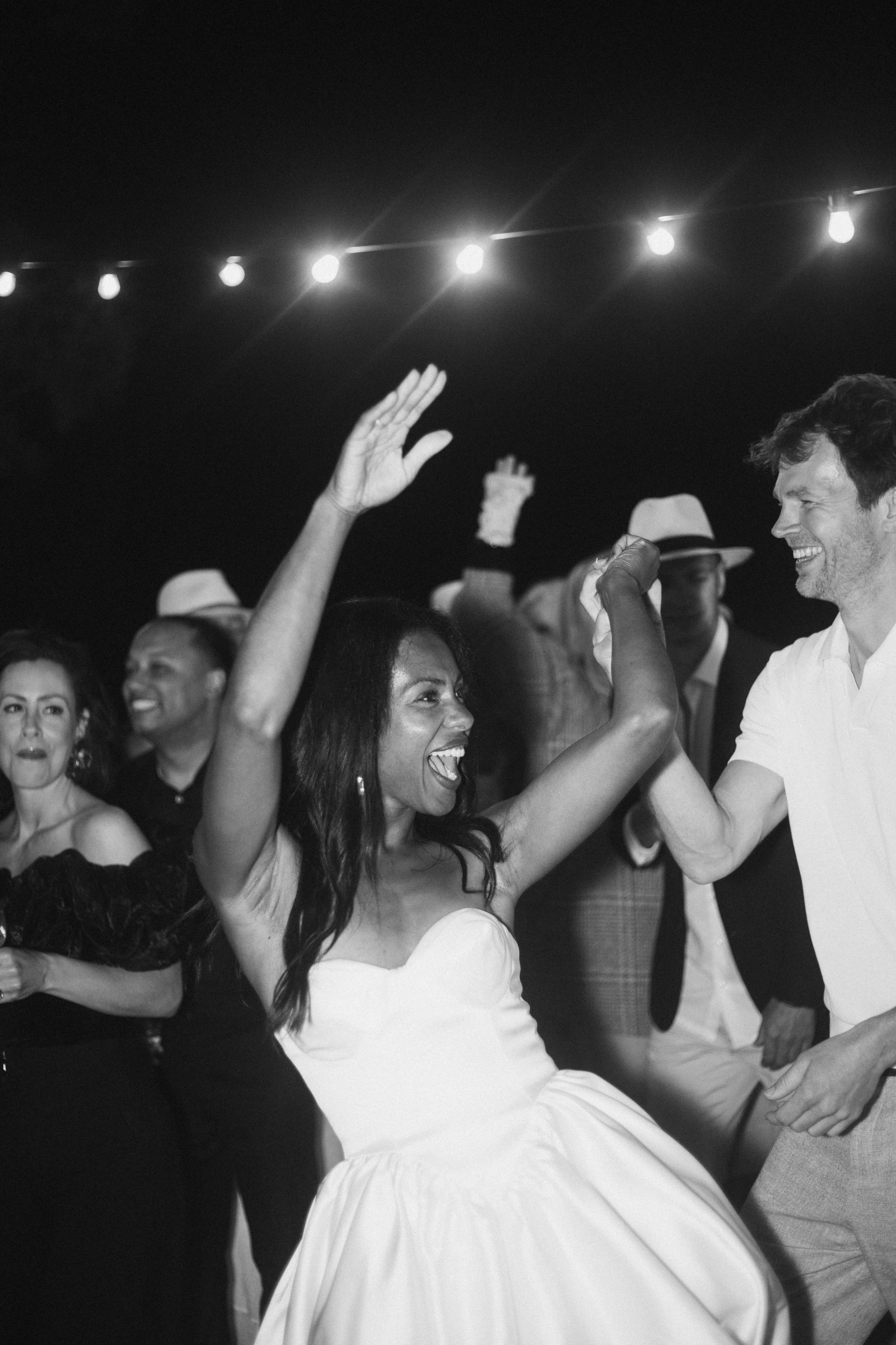 Un groupe de personnes dansant joyeusement lors d'une fête en plein air, une femme en robe blanche et un homme en chemise blanche dansent et sourient.
