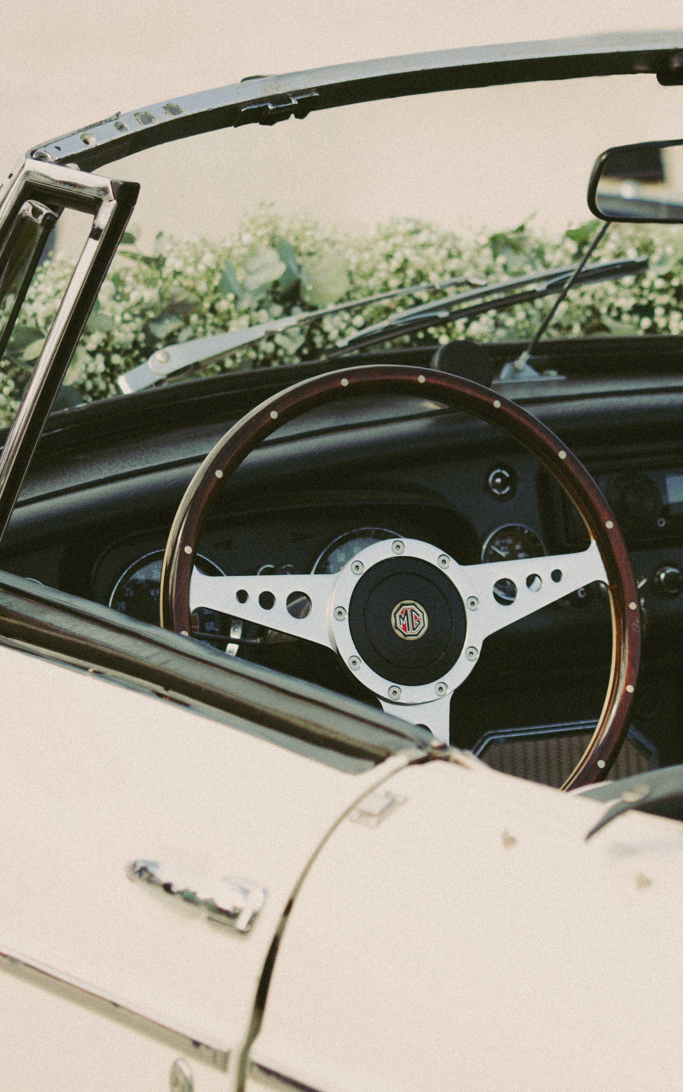 Intérieur d'une voiture classique avec un volant en bois et un logo MG, vue à travers le capot ouvert, avec des fleurs blanches en arrière-plan.