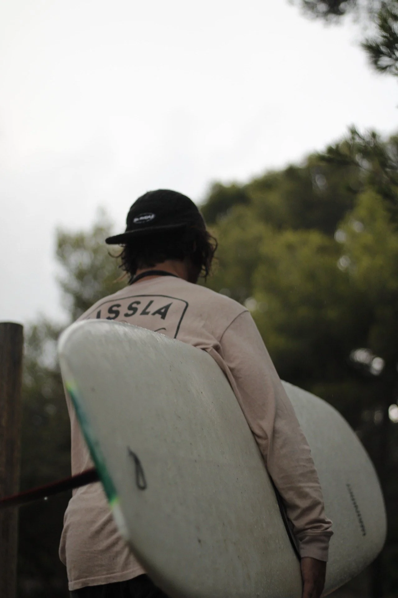 Une personne portant un chapeau noir et un t-shirt beige avec le mot "ISSLA" à l'arrière, tenant une planche de surf, vue de dos, dans un environnement naturel avec des arbres.