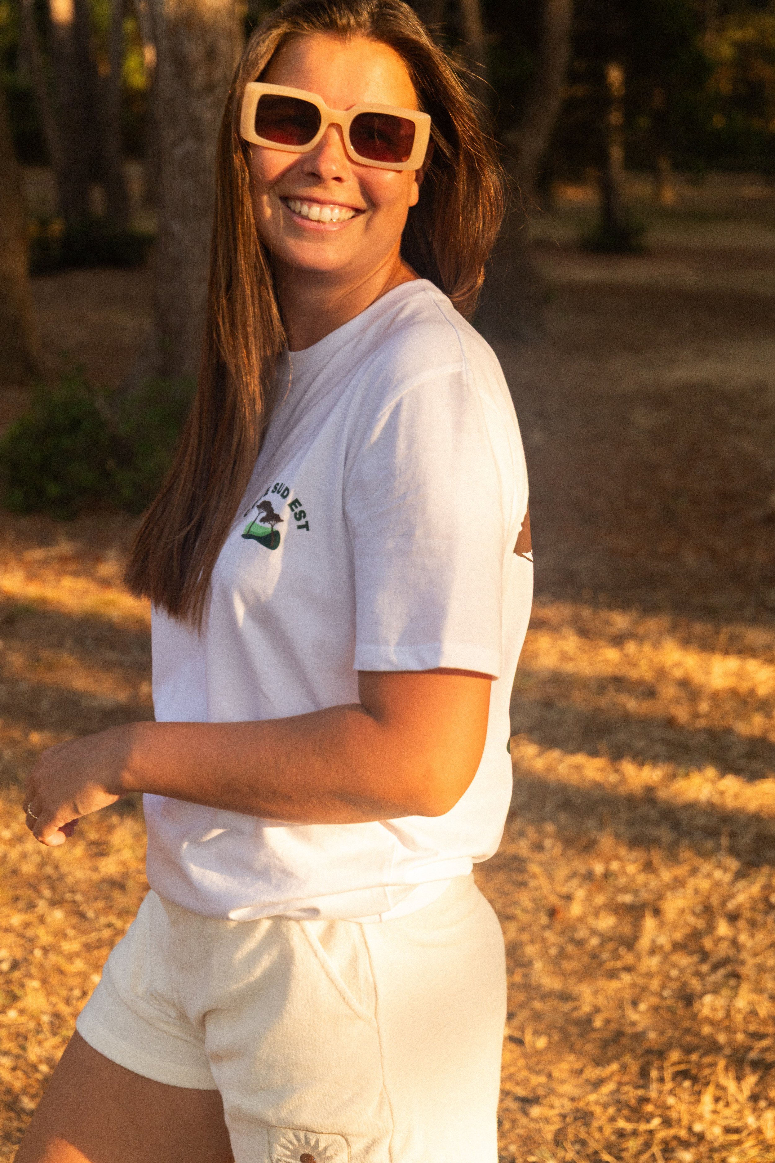 Femme souriante portant des lunettes de soleil, t-shirt blanc avec un logo, debout dans un parc en été, en pleine lumière de coucher de soleil.