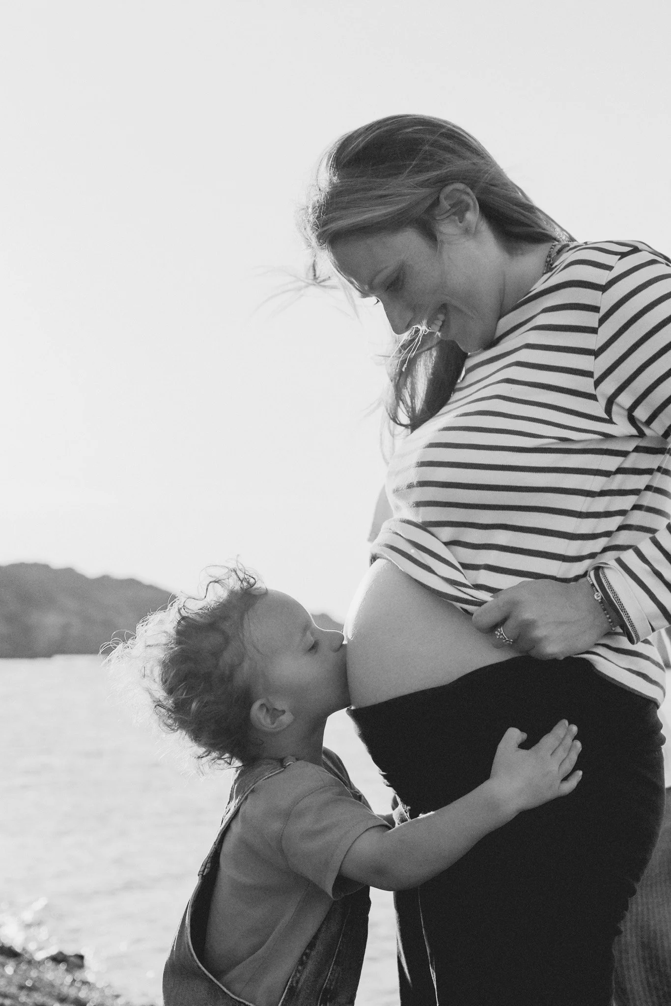 Une femme enceinte et un enfant qui l'embrasse sur le ventre near une rivière, en plein air, en noir et blanc.