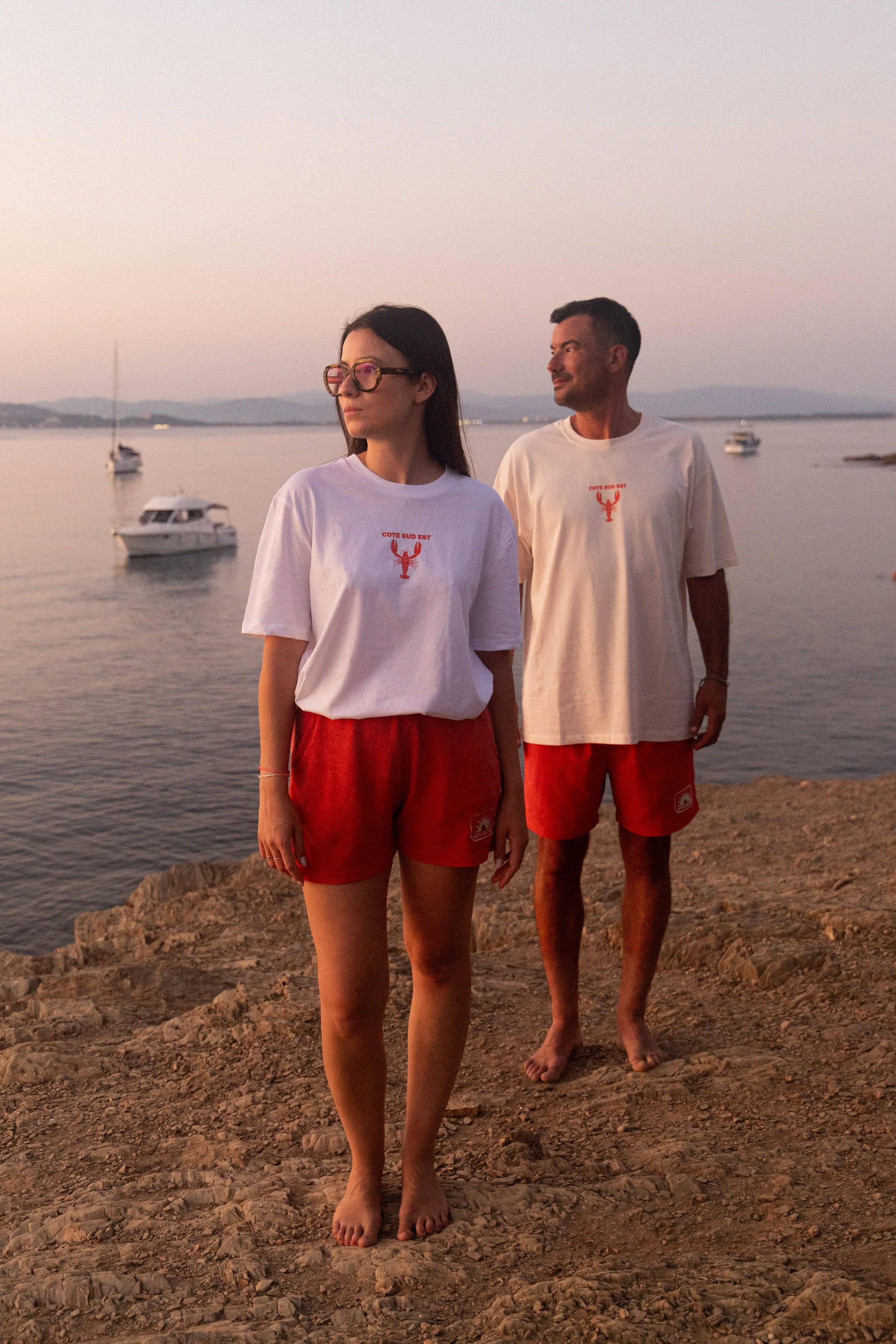 Deux personnes portant des T-shirts blancs et des shorts rouges se trouvent sur un rivage rocheux près d'un plan d'eau avec des bateaux en arrière-plan, au coucher du soleil.