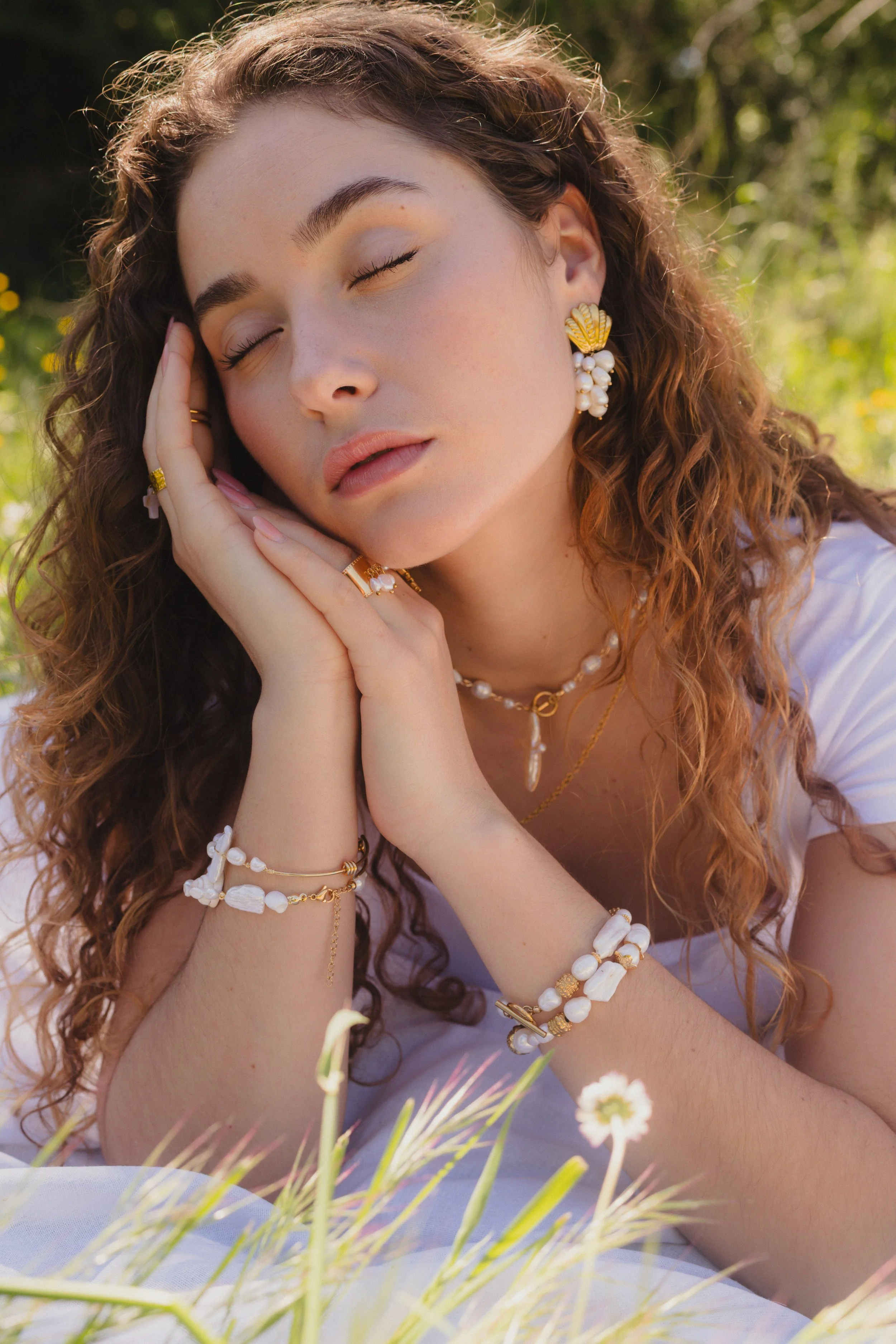 Jeune femme aux cheveux bouclés, portant des bijoux en coquillage et perles, allongée dans un champ ensoleillé avec des fleurs.