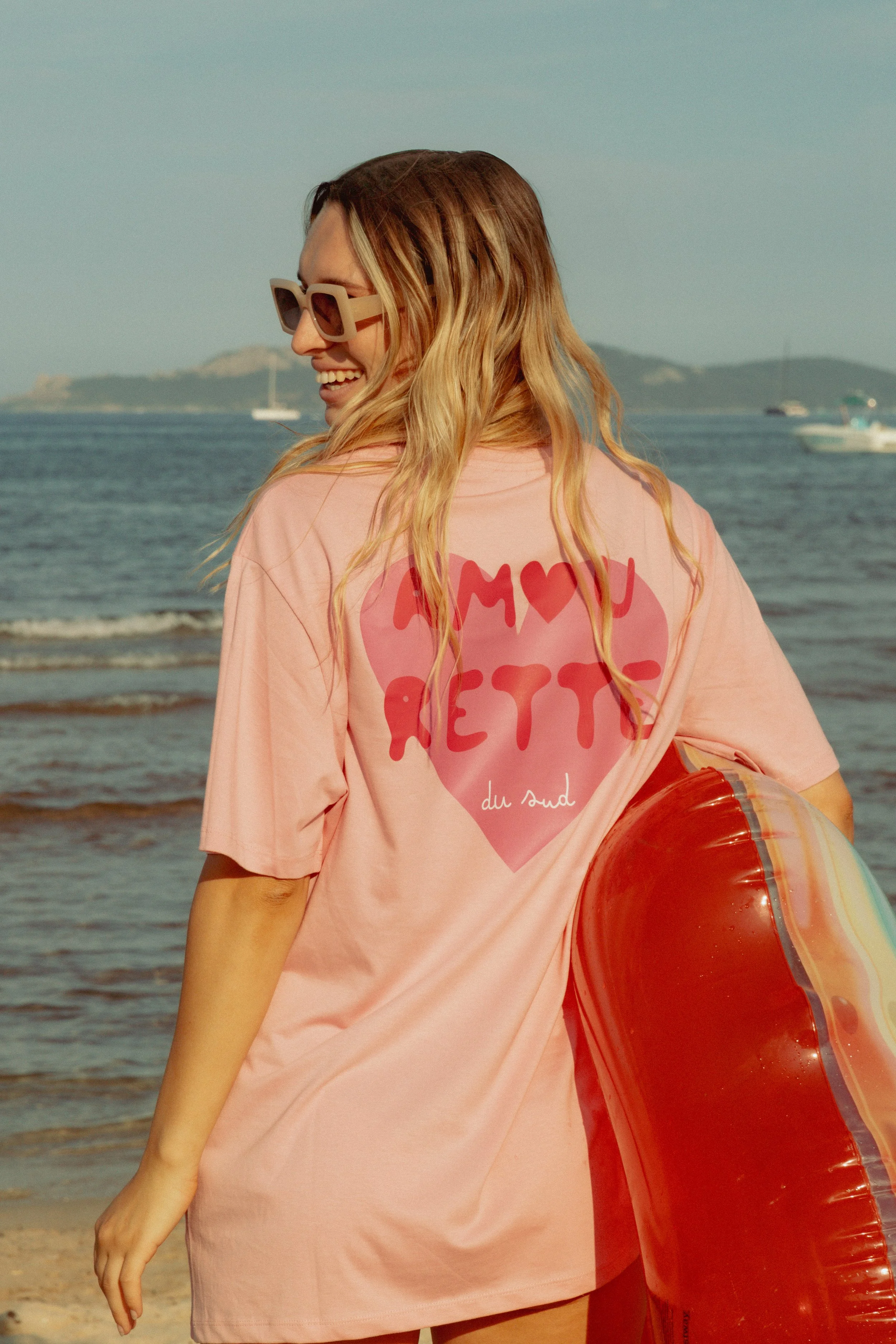 Une femme souriante portant des lunettes de soleil, portant un t-shirt rose avec un message en français, tenant une bouée de plage sur la plage avec la mer en arrière-plan.