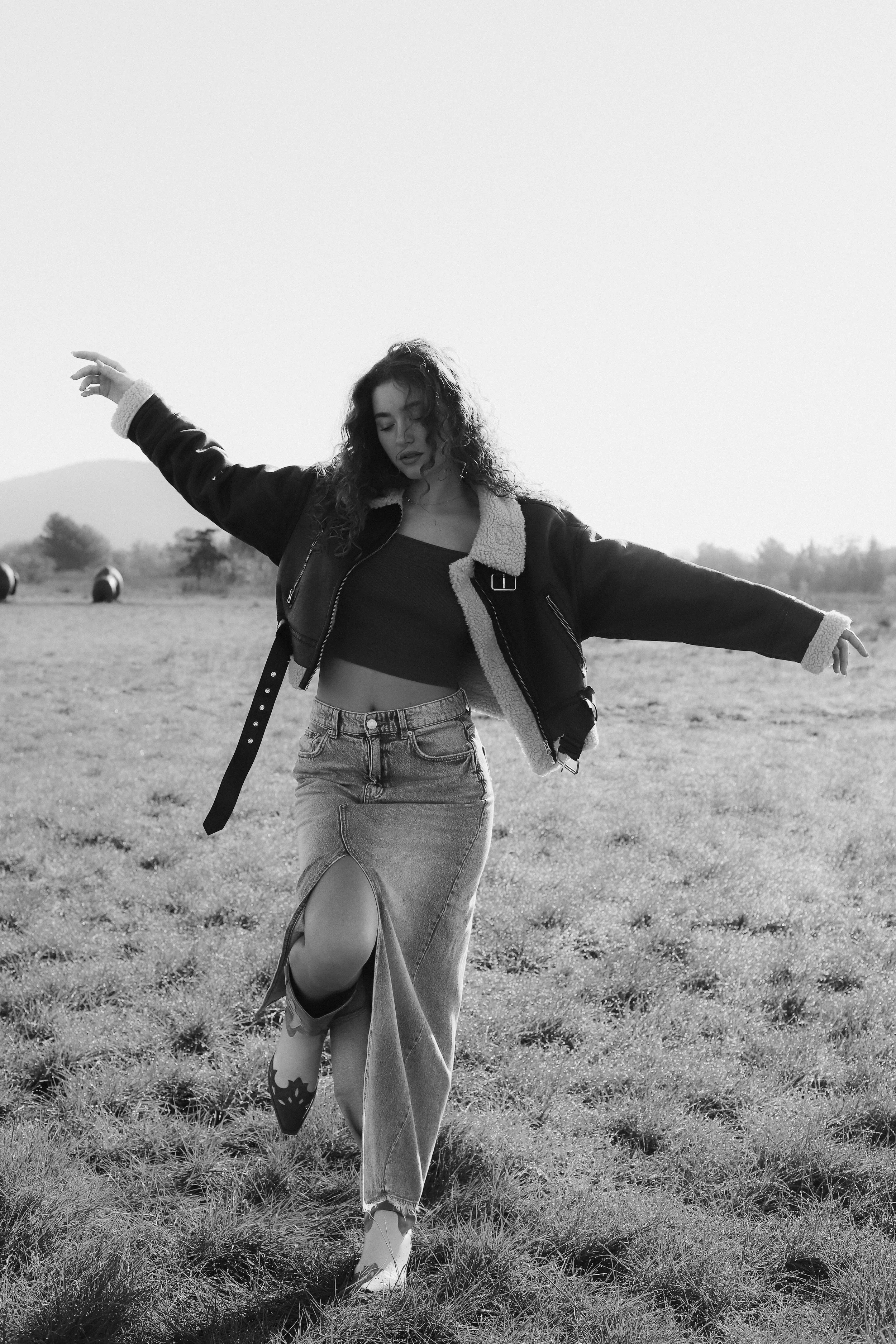 Une jeune femme marche dans un champ, portant un jean déchiré, un haut court et une veste en cuir. Elle a les bras étendus et semble danser ou profiter du moment.