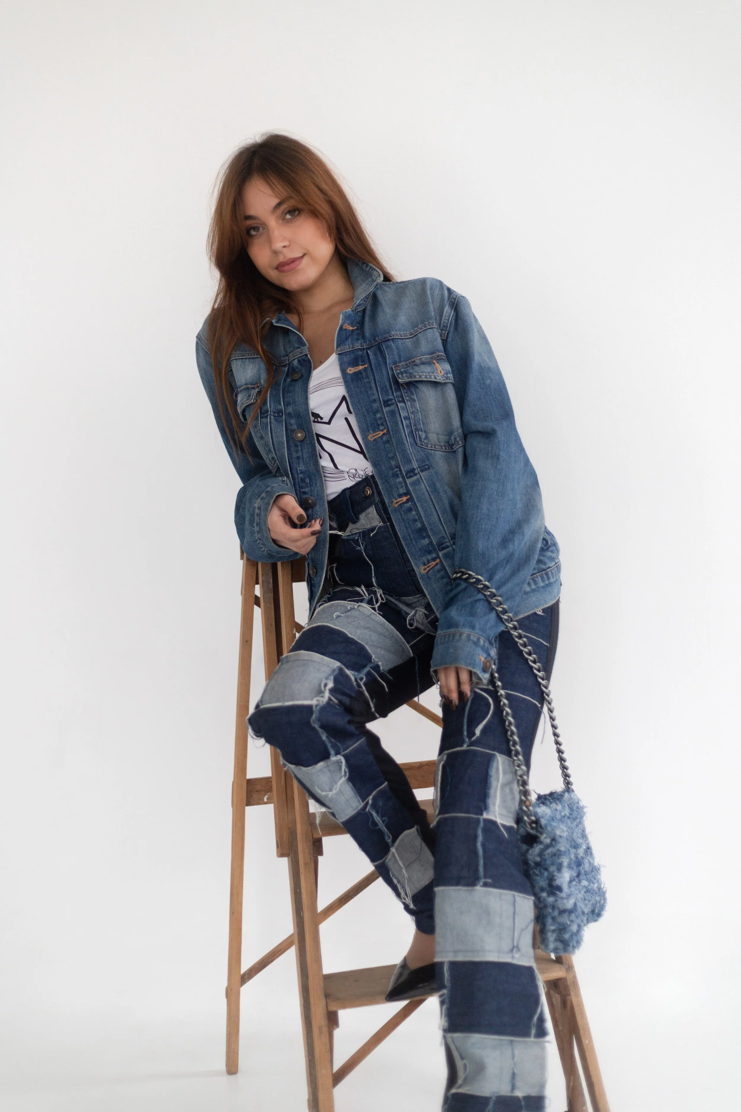 Jeune femme aux cheveux bruns, portant un jean déchiré et une veste en jean, assise sur un escabeau en bois, tenant un téléphone portable dans une main et un sac à main en fausse fourrure bleue, dans un studio avec un fond blanc.