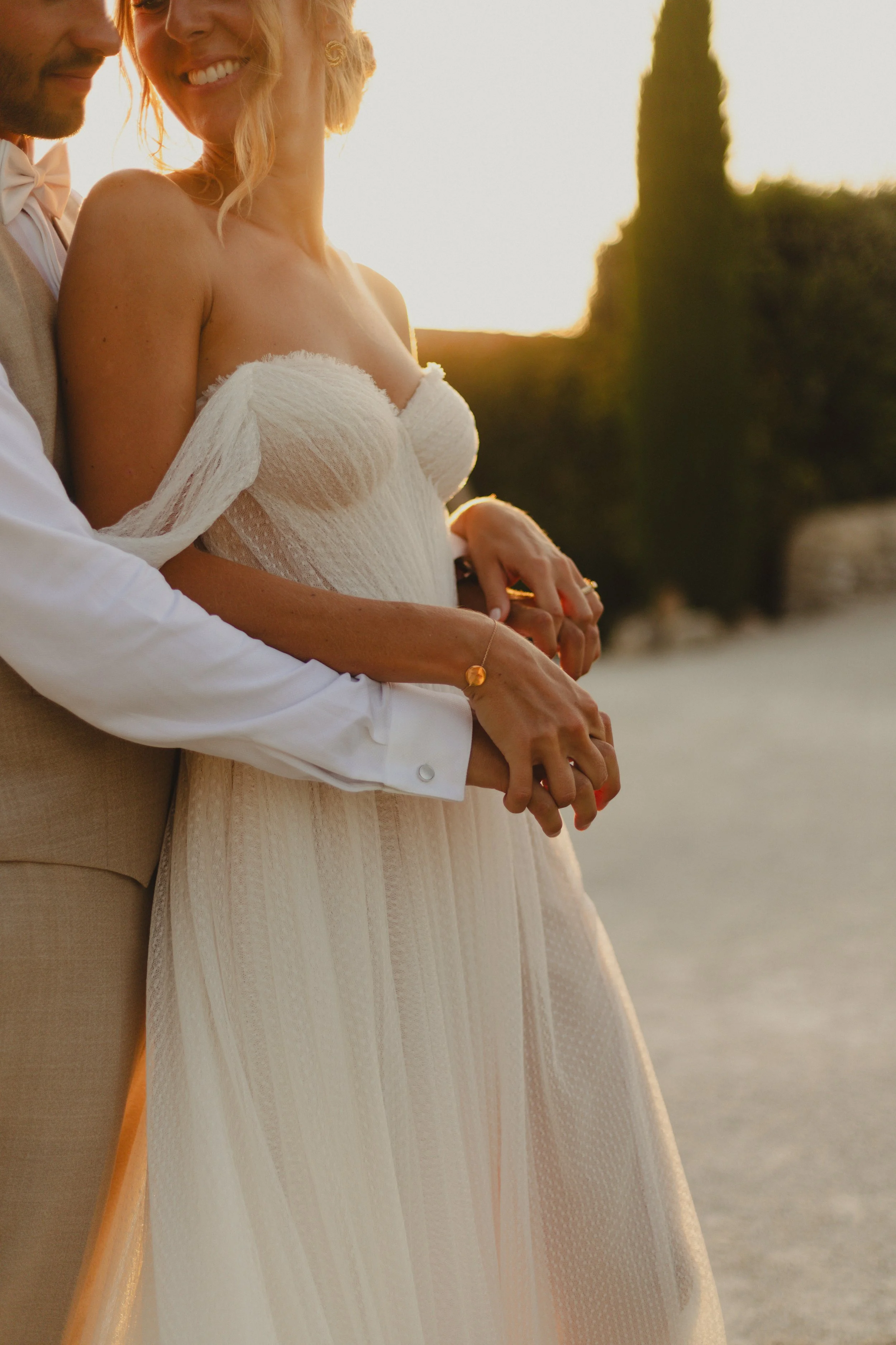 Un couple en tenue de mariage, l'homme en costume beige et la femme en robe blanche, se tenant par la main lors d'une séance photo au coucher du soleil dans un lieu extérieur.