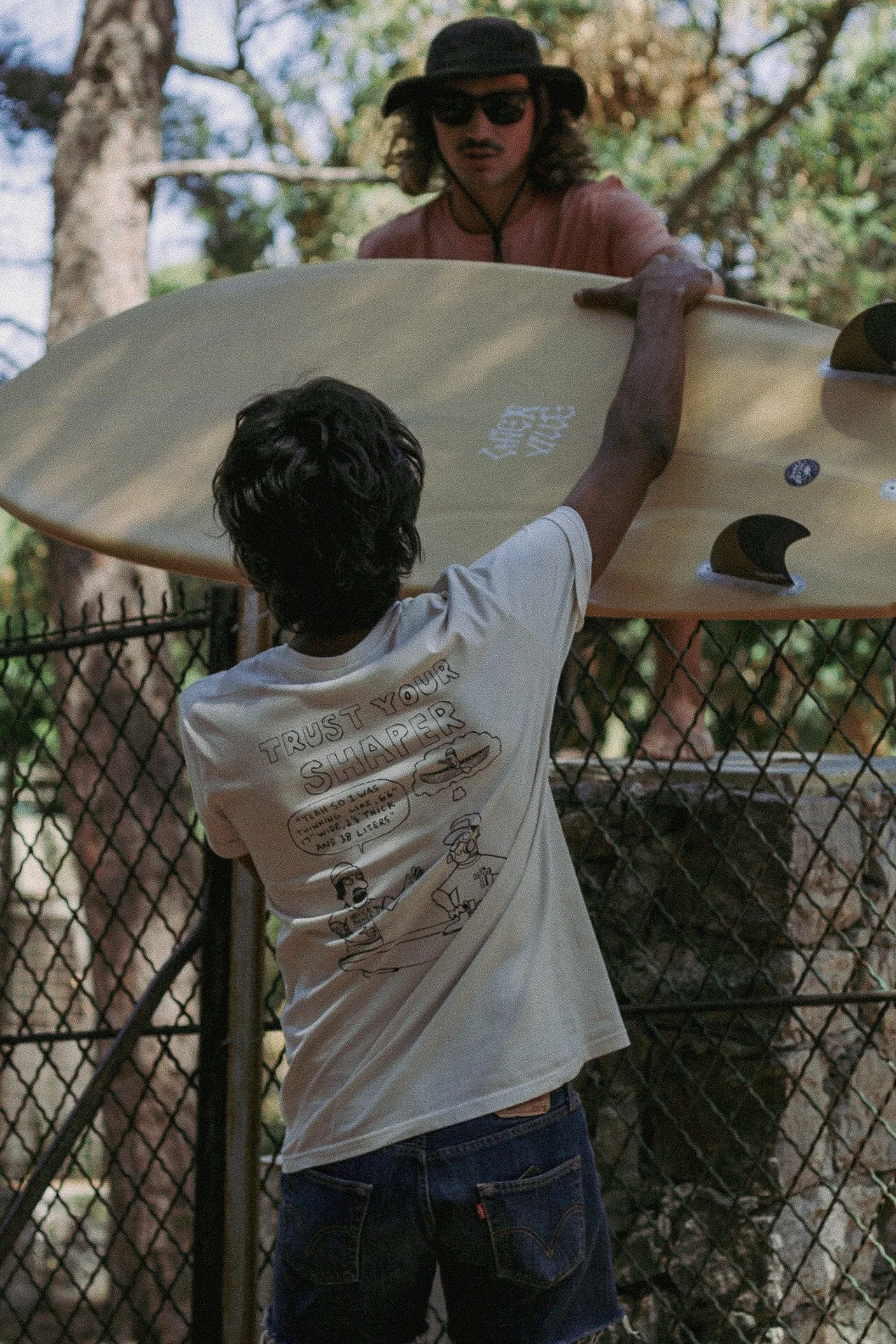 Jeune garçon remettant une planche de surf à un homme portant un chapeau et des lunettes de soleil, en extérieur près d'une clôture en métal, avec des arbres en arrière-plan.