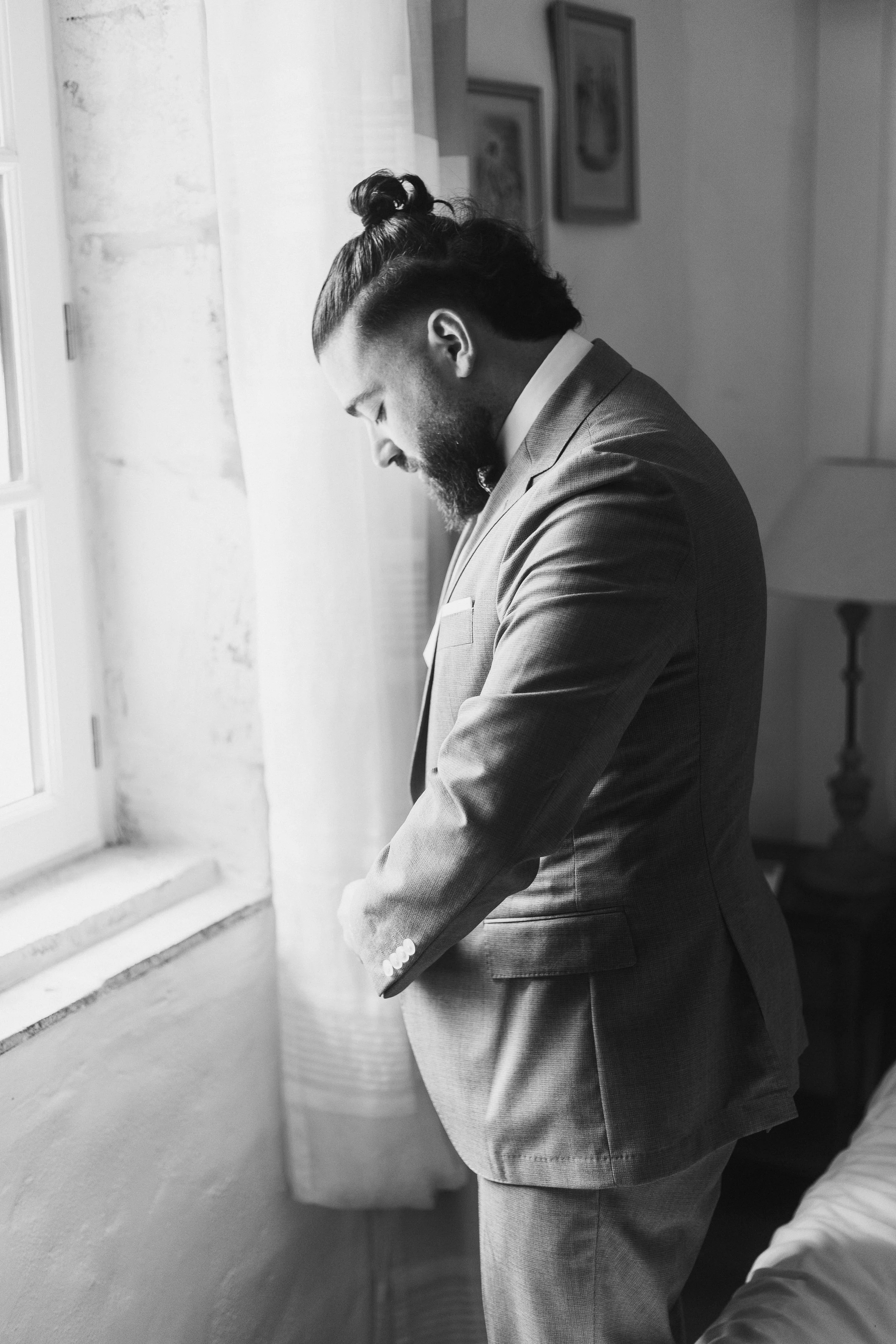Un homme portant un costume ajuste, debout près d'une fenêtre, regardant vers le bas avec la tête inclinée.
