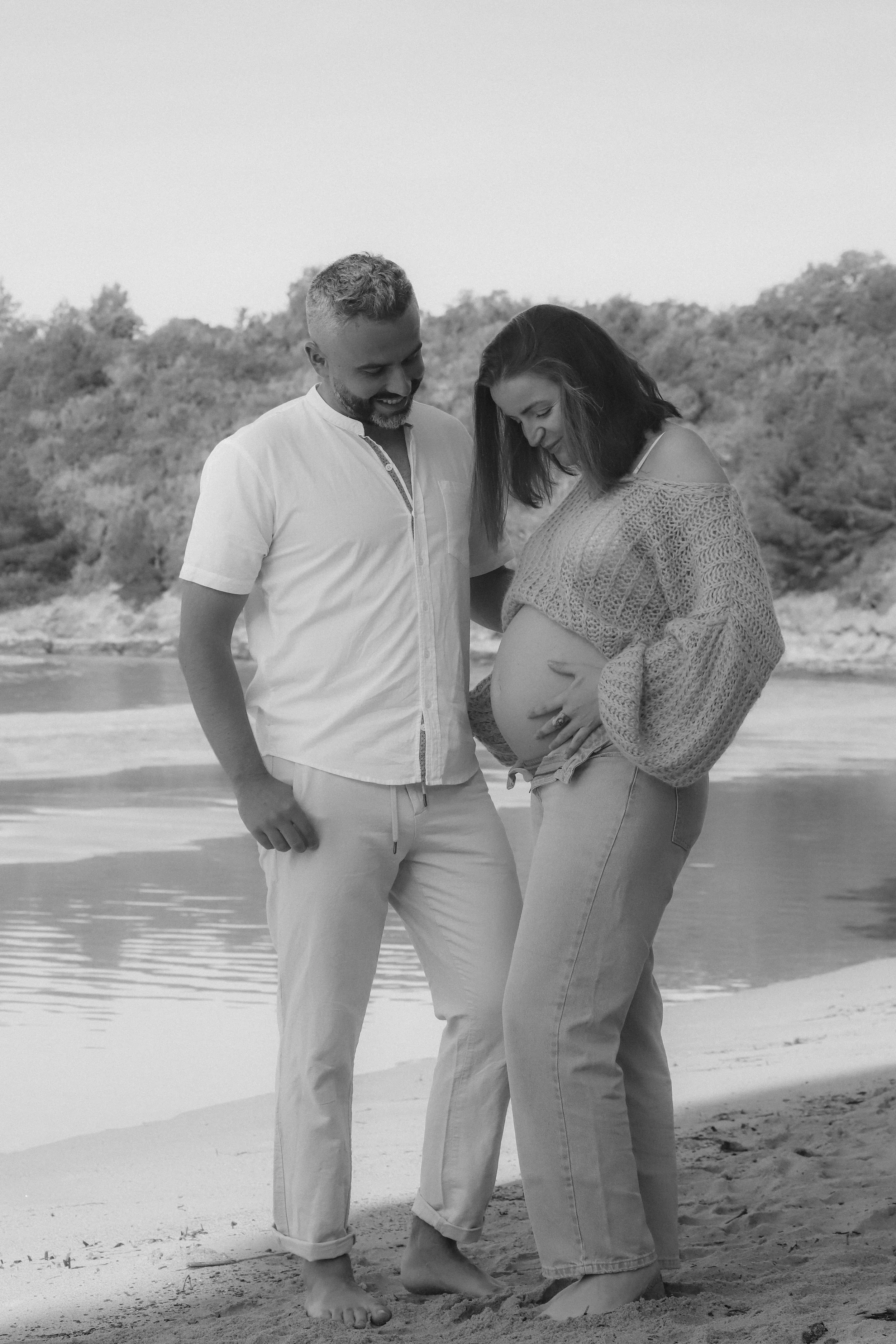 Un couple souriant, le ventre de la femme enceinte visible, se tenant par la main sur la plage.