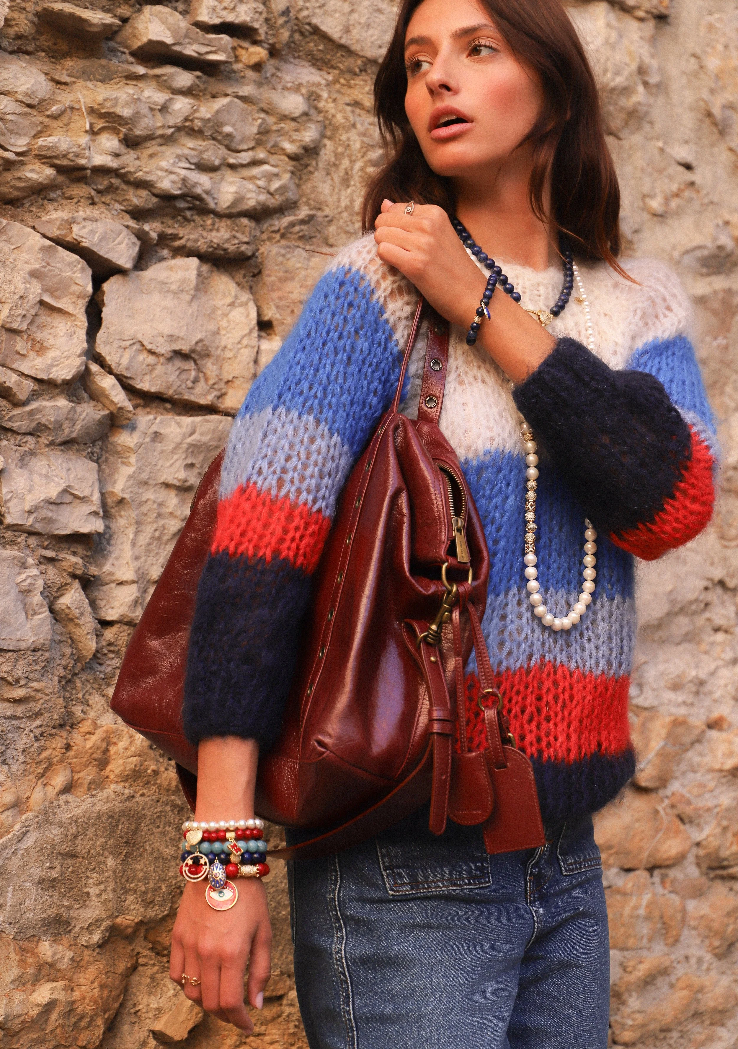 Une femme portant un pull coloré en maille, un sac en cuir marron, des bijoux en perles et plusieurs bracelets, se tient devant un mur en pierre.
