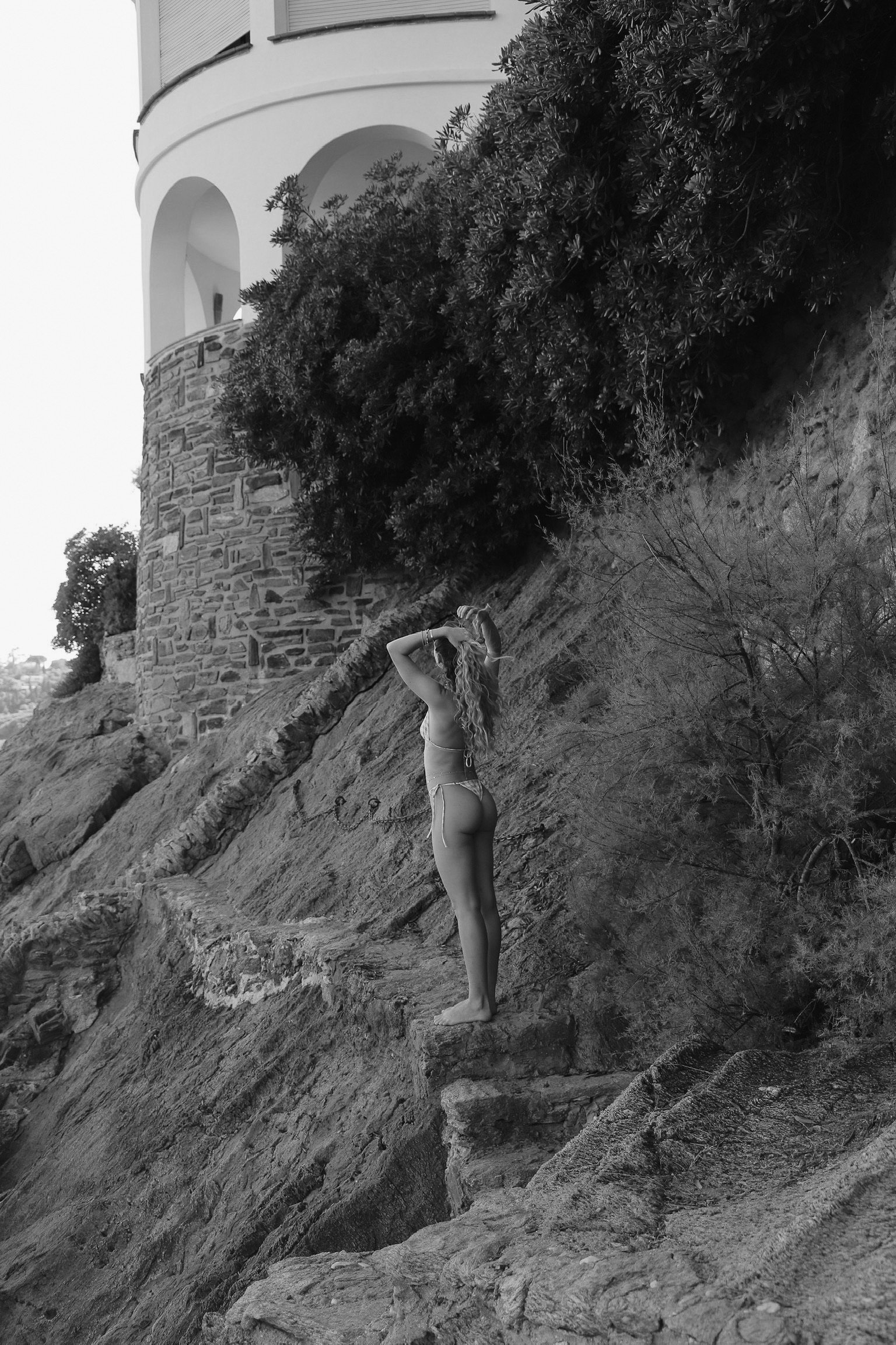 Une femme en maillot de bain se tient pieds nus sur des rochers, regardant vers le haut, avec une maison en pierre en arrière-plan. La photo est en noir et blanc.