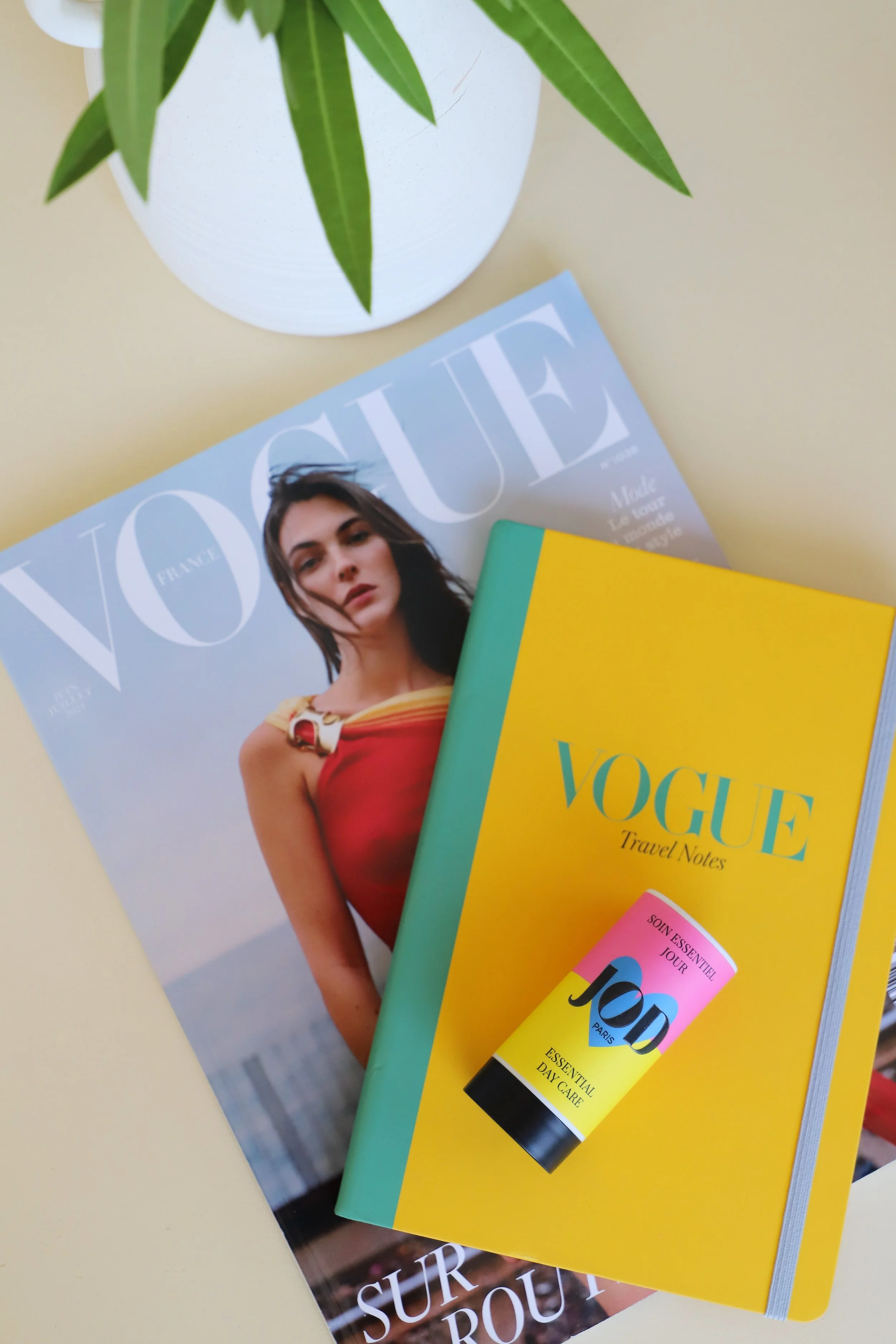 Une magazine Vogue, un carnet VOGUE Travel Notes, un tube de soin JODY, un pot de fleurs vert avec des feuilles vertes dans un vase blanc