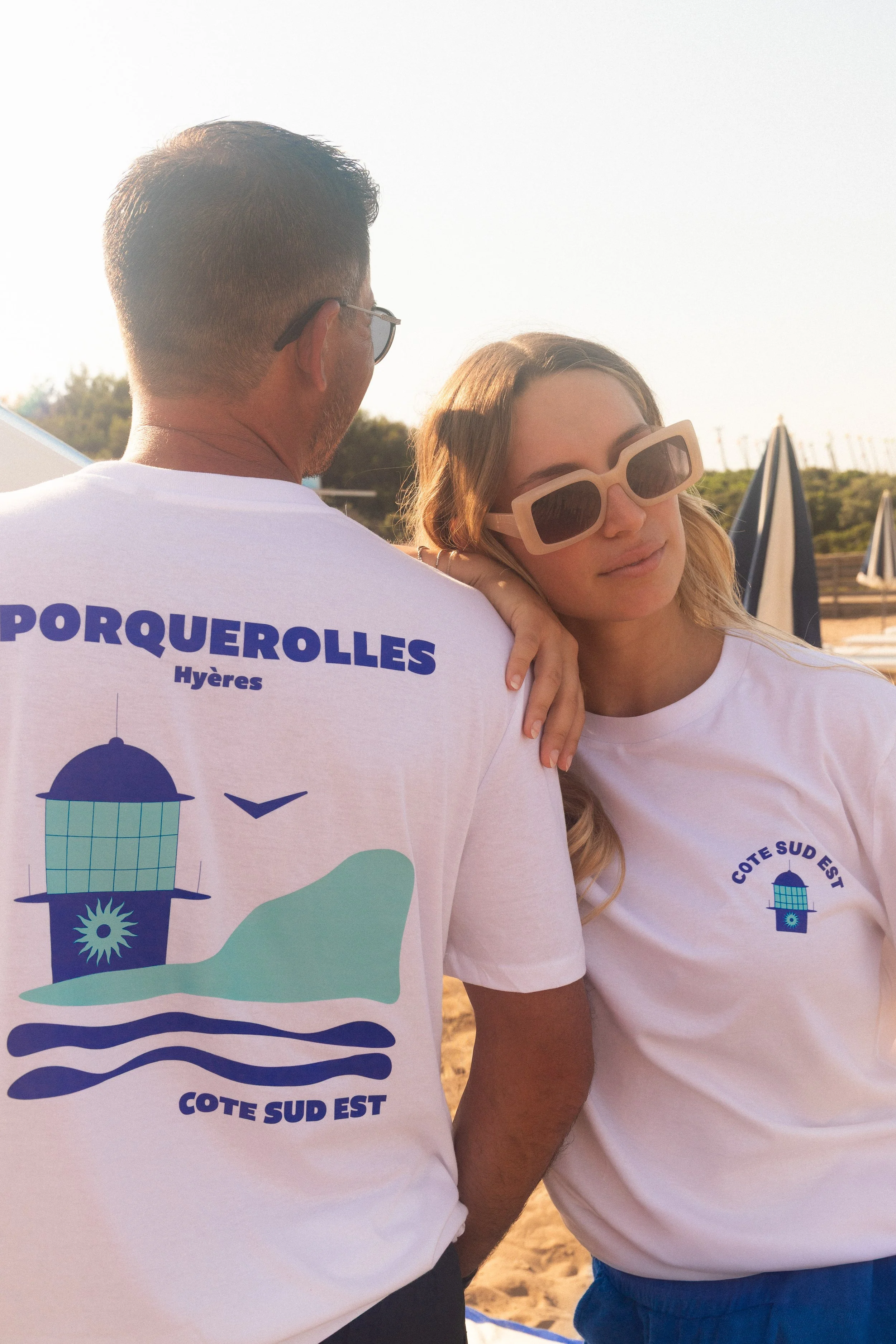 Deux personnes portent des t-shirts blancs avec des motifs et textes liés à la Côte Sud Est, dans un décor en plein air avec des parasols et un ciel ensoleillé.