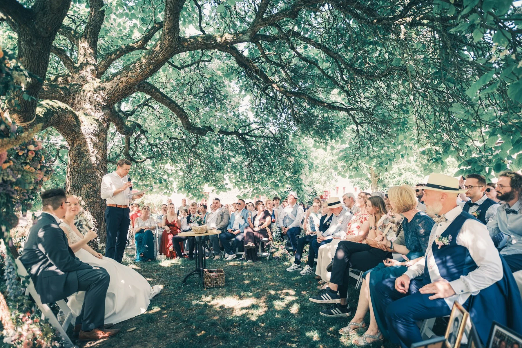 Cérémonie laïque en plein air sous de grands arbres, invités assis en rangs autour du couple – mariage en Belgique