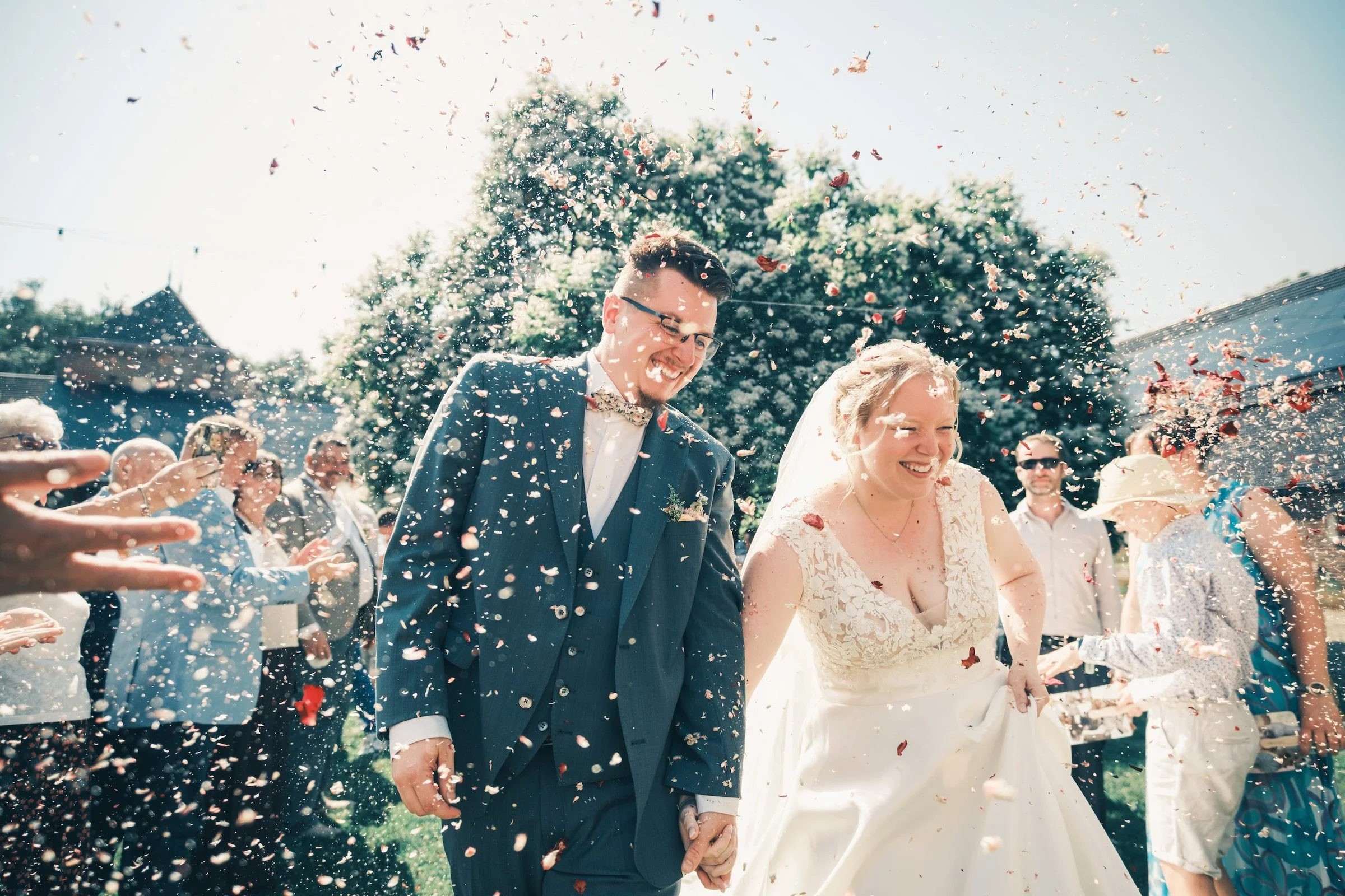 Mariés qui rient aux éclats sous une pluie de pétales à la sortie de la cérémonie – reportage de mariage À Tantôt