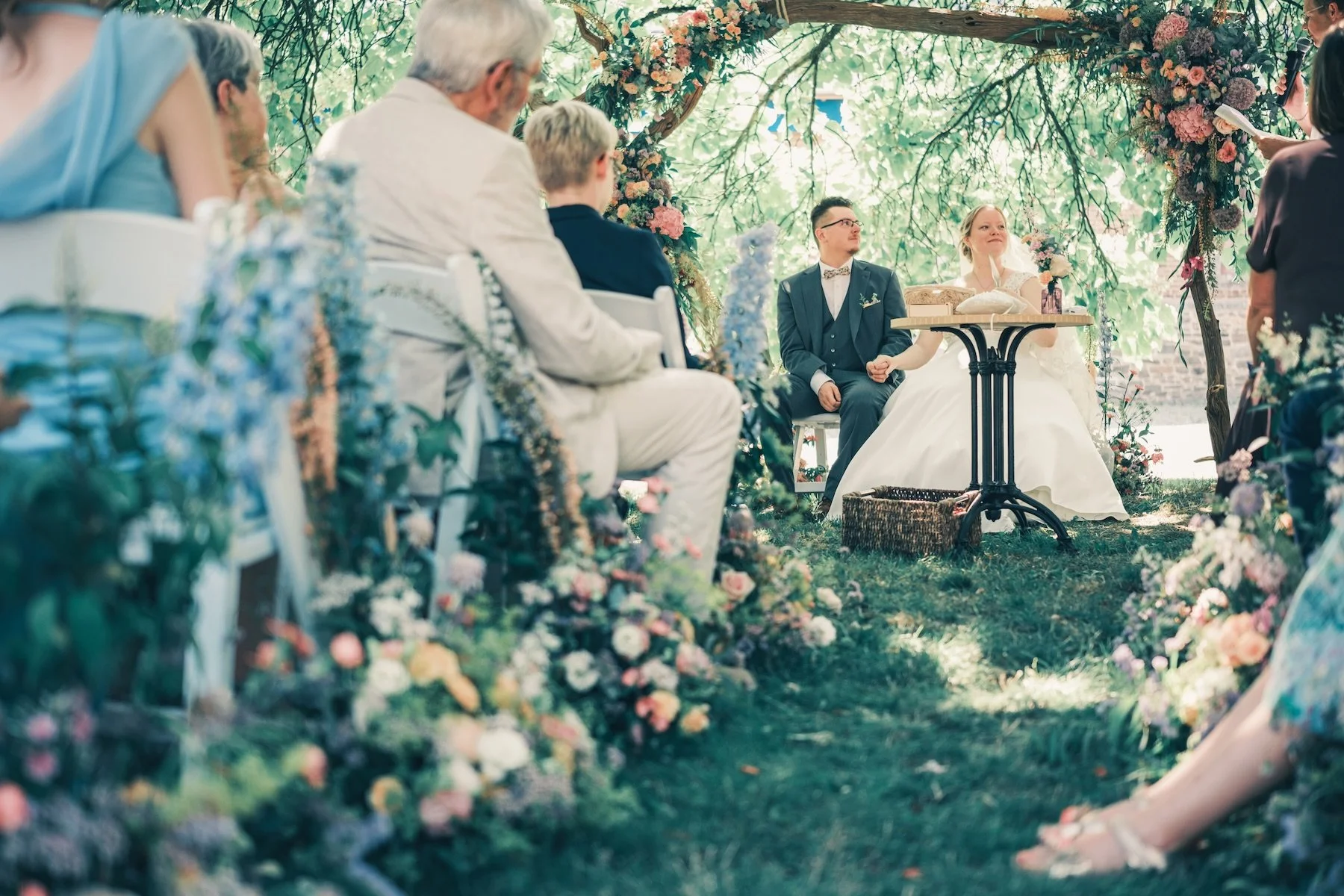Cérémonie laïque sous une arche fleurie, les mariés assis à une petite table ronde face à leurs invités