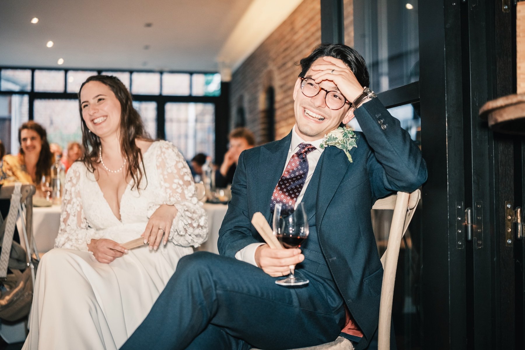 Mariés qui rient à leur table pendant le repas de mariage, le marié tenant un verre de vin rouge