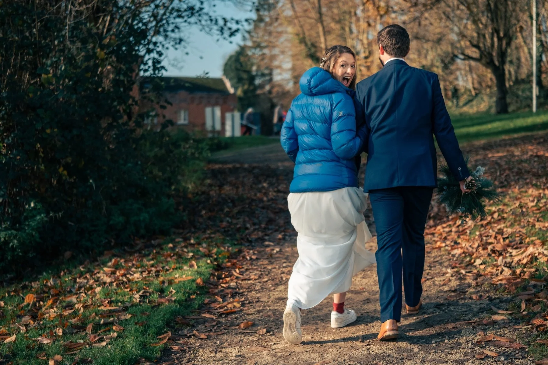 Mariés de dos qui marchent sur un chemin d'automne, la mariée en doudoune bleue sur sa robe – mariage d'automne