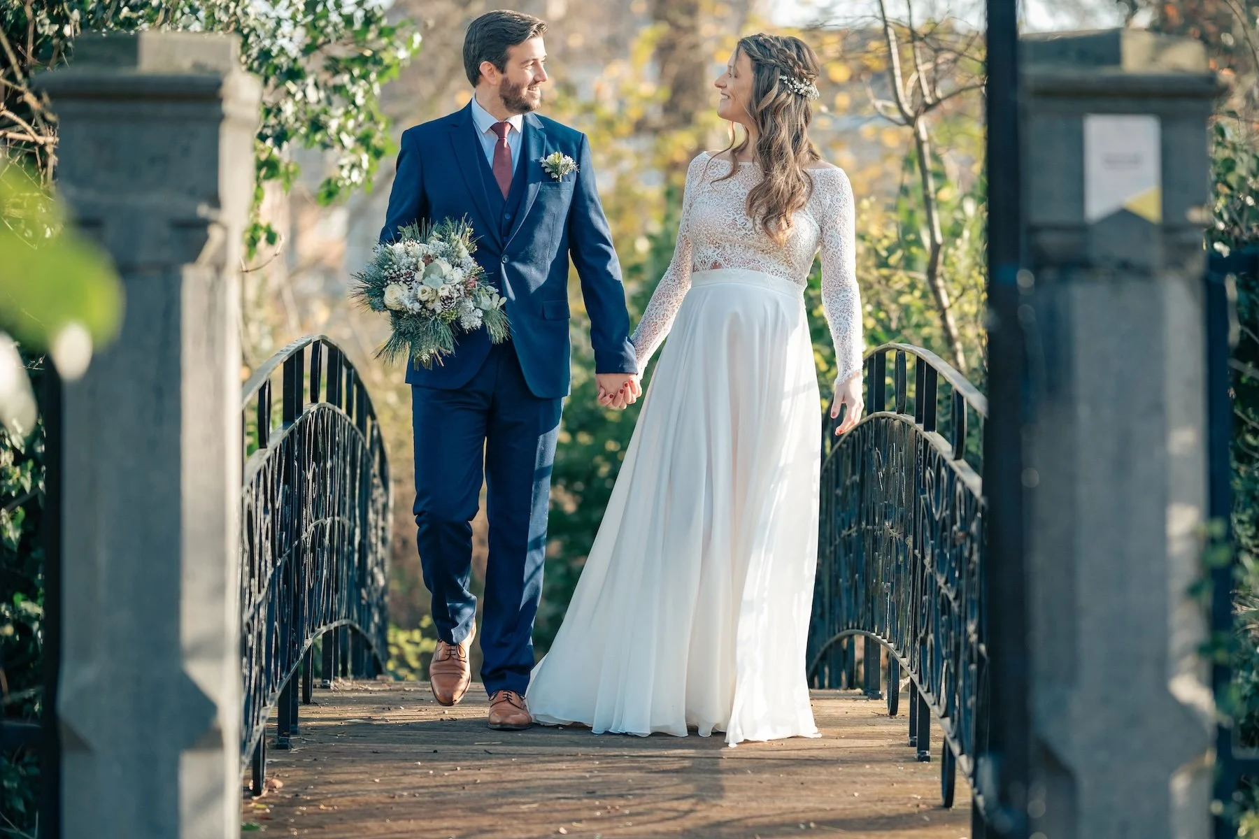 Mariés main dans la main sur un pont en fer forgé en automne, bouquet de fleurs – séance couple de mariage en Belgique