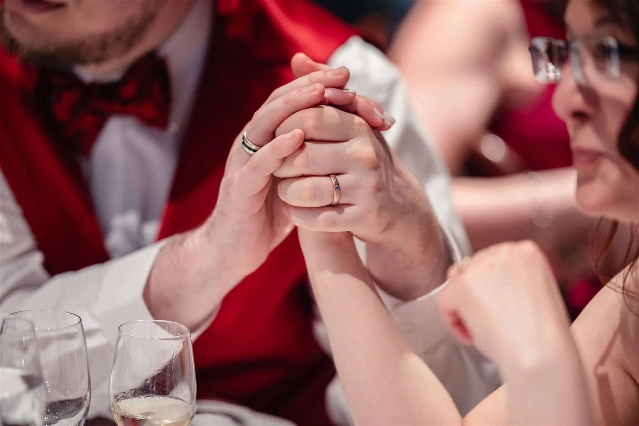 Gros plan des alliances entre les mariés, gros plan sur leurs mains et leurs bagues, avec des verres de vin en arrière-plan.