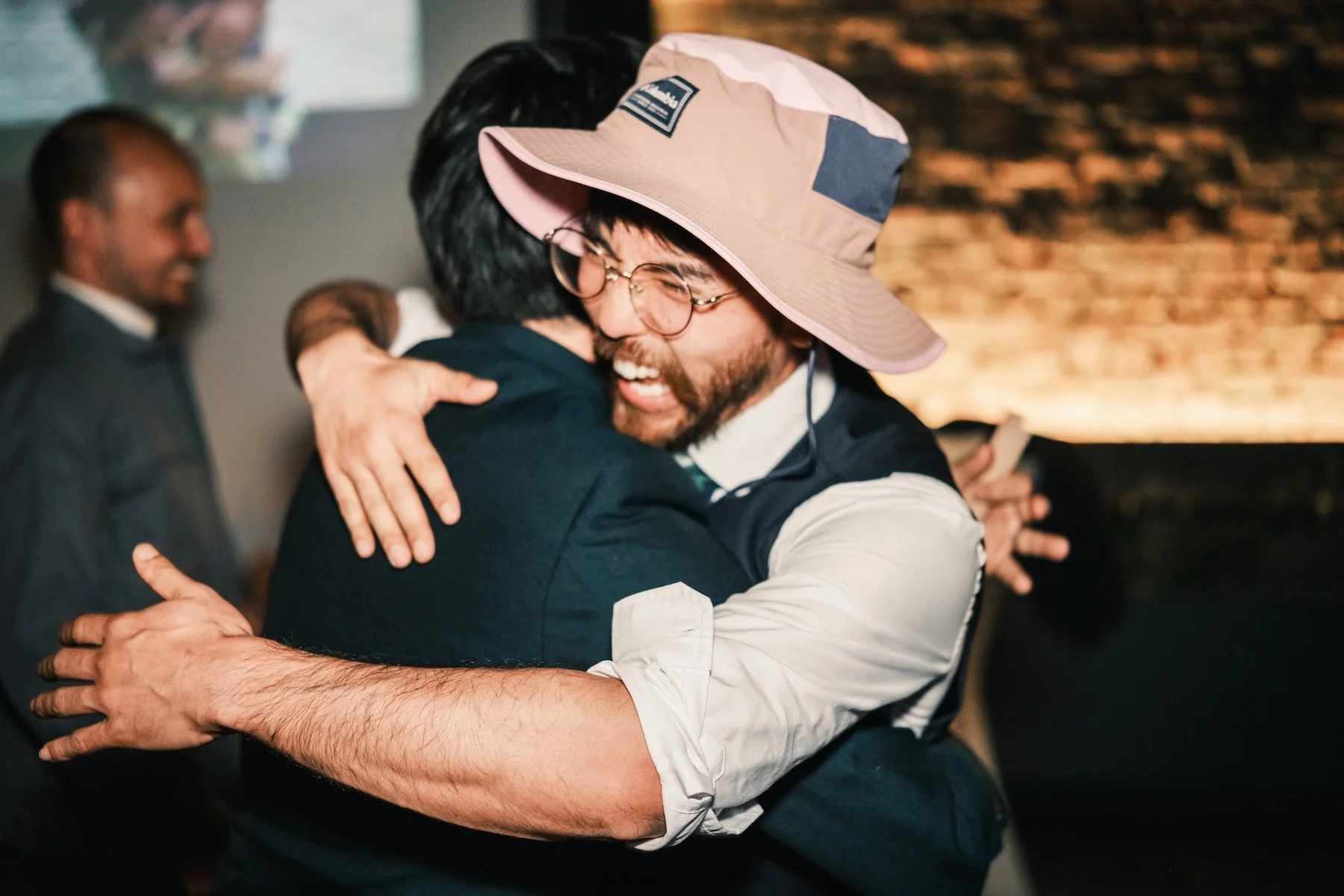 Invité barbu en chapeau rose qui rit aux éclats en serrant un ami dans ses bras pendant la fête de mariage