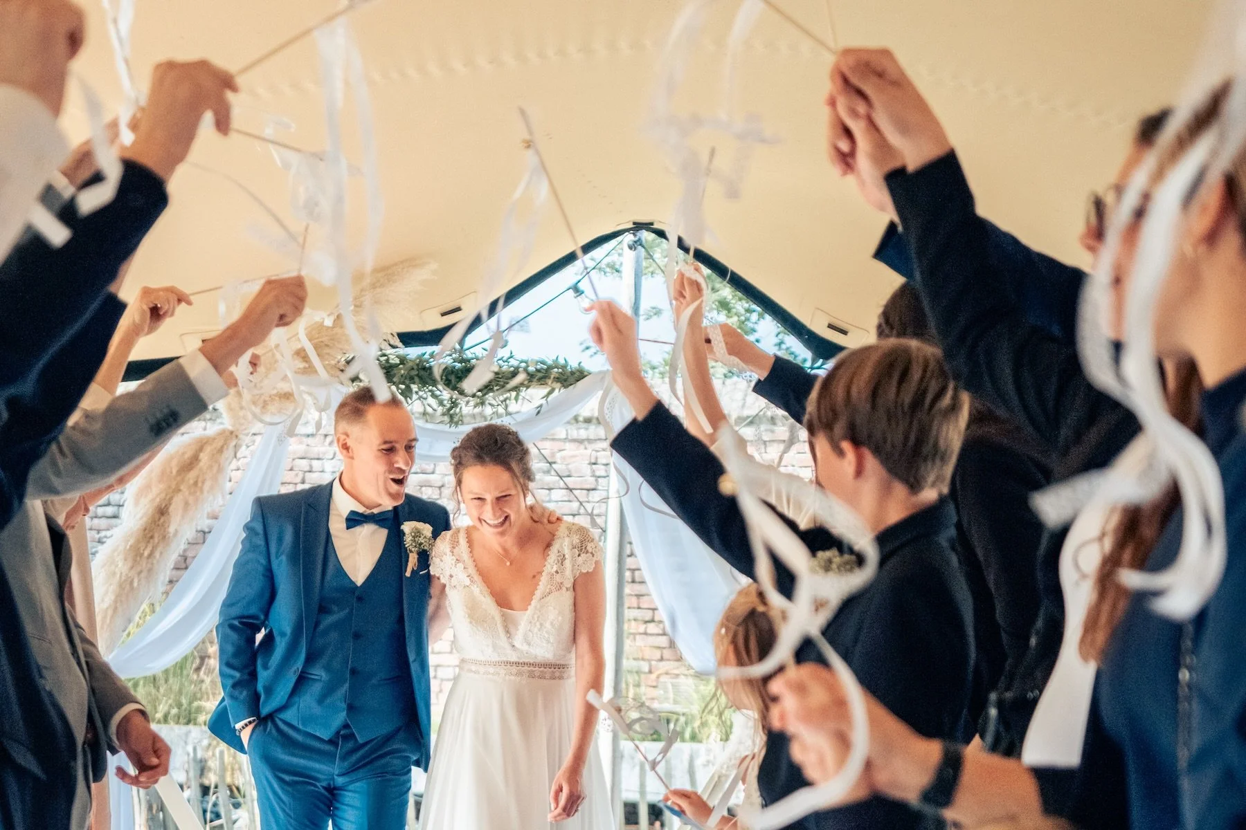 Mariés qui sortent de la cérémonie sous une arche de rubans tenue par leurs invités, sous une tente décorée de pampas