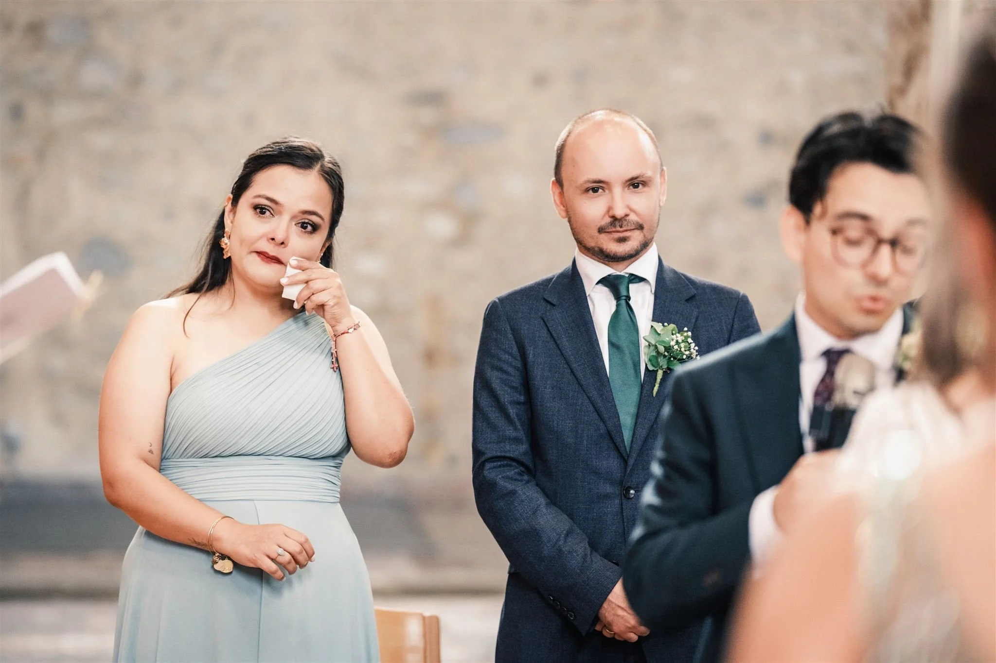 Témoin émue essuyant une larme pendant la cérémonie de mariage, avec les mariés et l’officiant en arrière-plan.