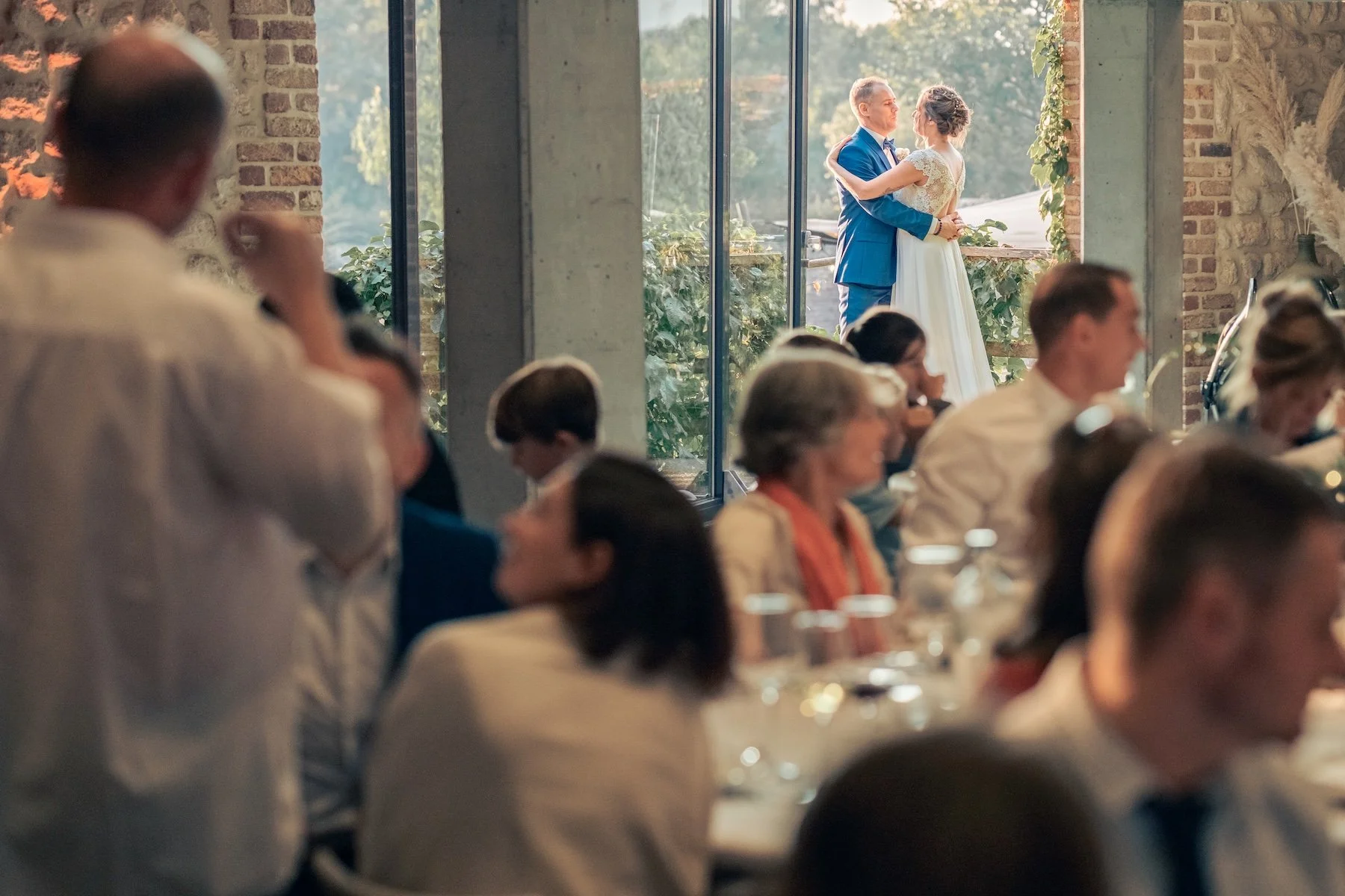 Dîner de mariage : les mariés dansent au fond de la salle devant une grande baie vitrée, au milieu des invités attablés