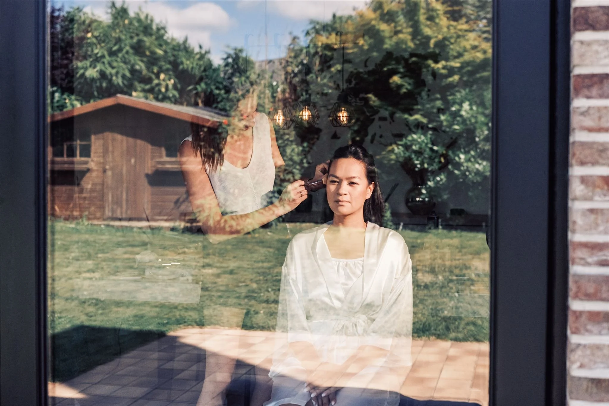 Mariée en peignoir blanc pendant son maquillage, reflets du jardin dans la vitre – préparatifs de mariage