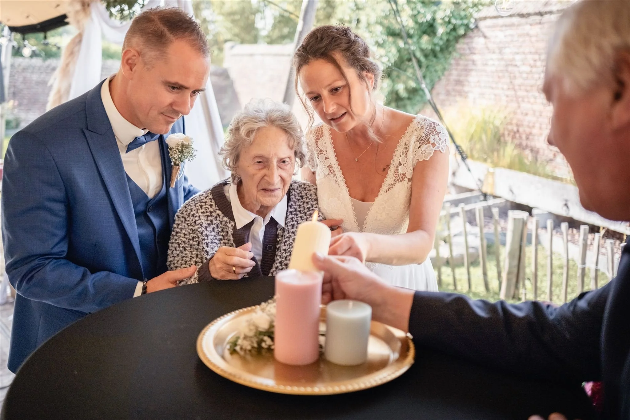 eune couple allumant une bougie avec leur grand-mère lors d’un rituel symbolique pendant leur mariage en extérieur.