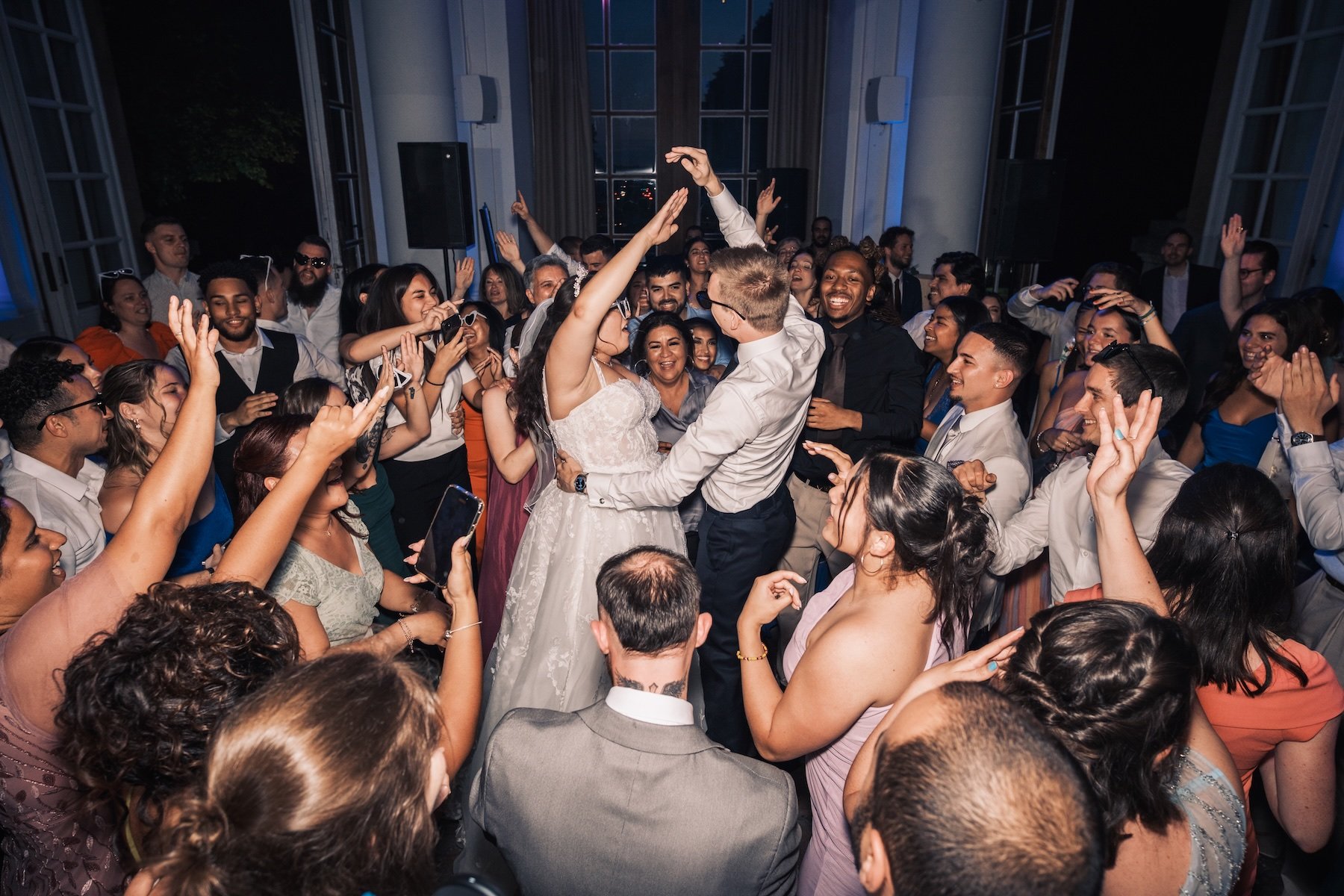 Mariés entourés d'invités aux bras levés sur la piste de danse, ambiance de fête dans une salle à grandes baies