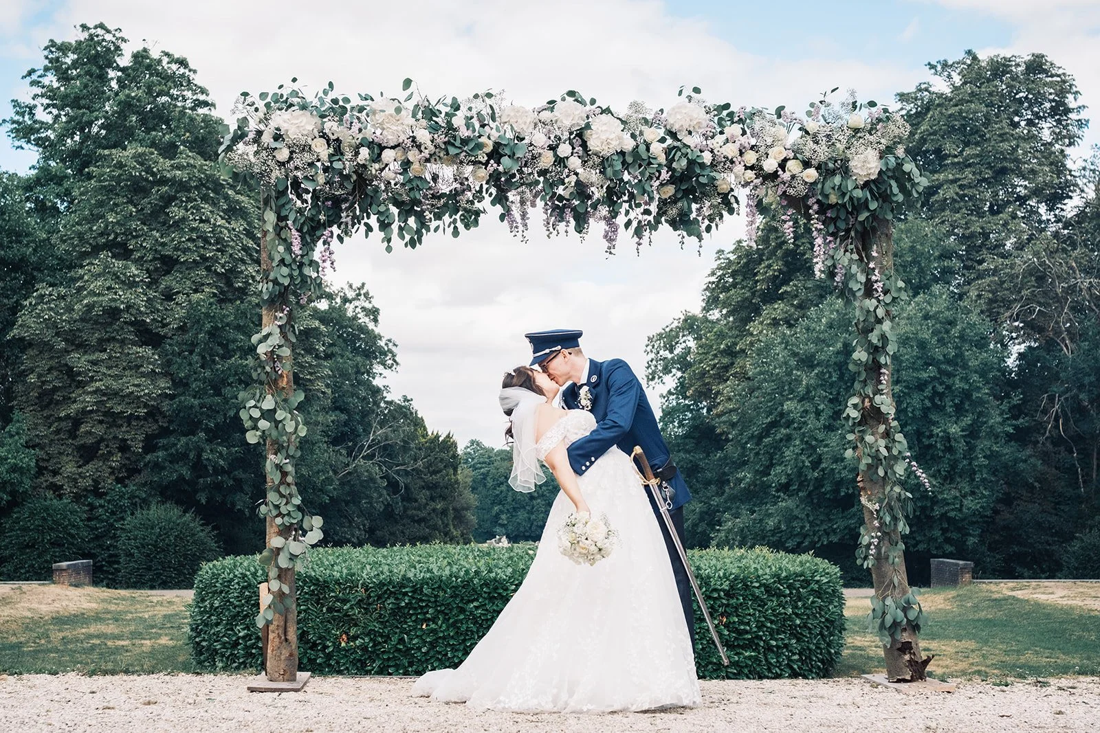 Baiser des mariés sous une arche fleurie en plein air, entourés de verdure, lors d’une séance photo romantique et naturelle.