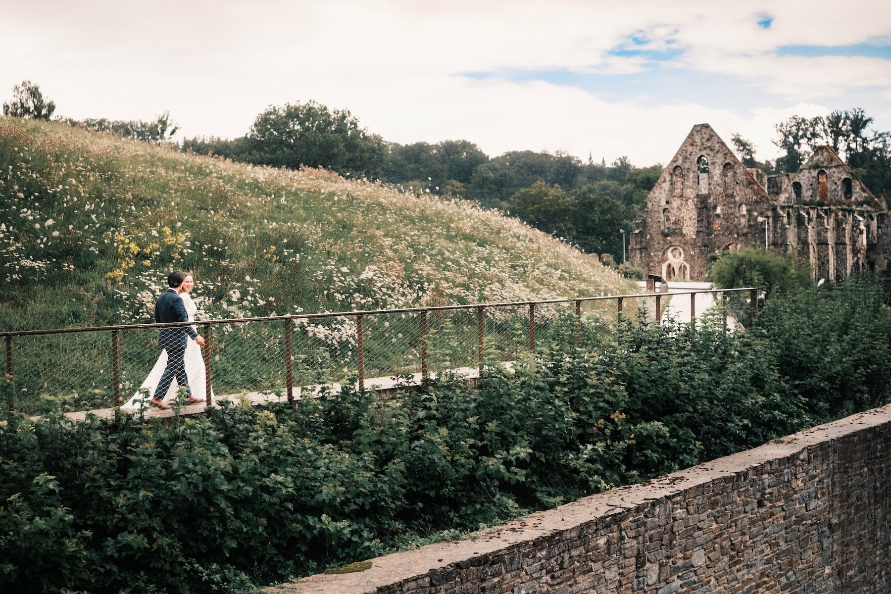 Mariés qui marchent au pied d'une colline fleurie, devant les ruines d'une abbaye – séance couple de mariage en Belgique