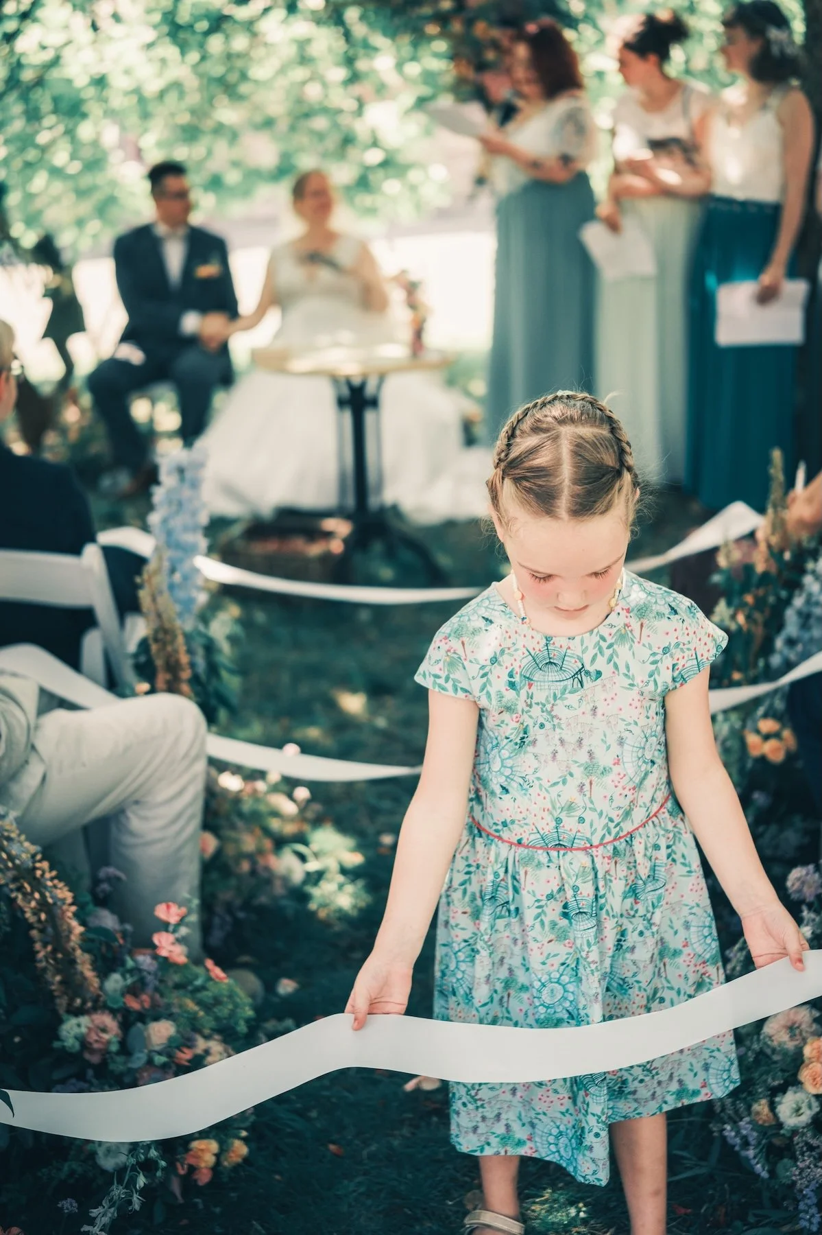 Petite fille en robe fleurie tenant un ruban lors d'une cérémonie laïque dans un jardin fleuri, mariés en arrière-plan