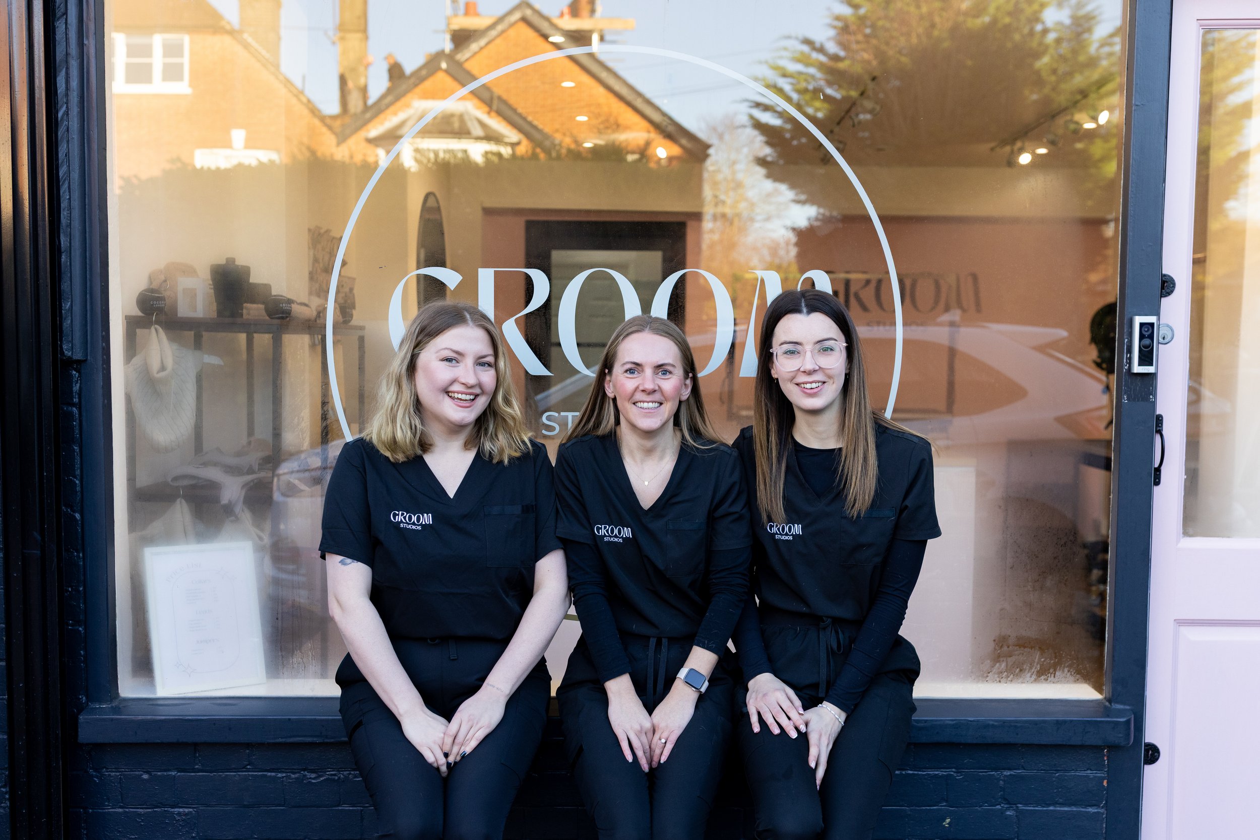 Three women wearing black uniforms sitting in front of a beauty salon window with a sign that says 'GROOM' and a reflection of houses and trees.