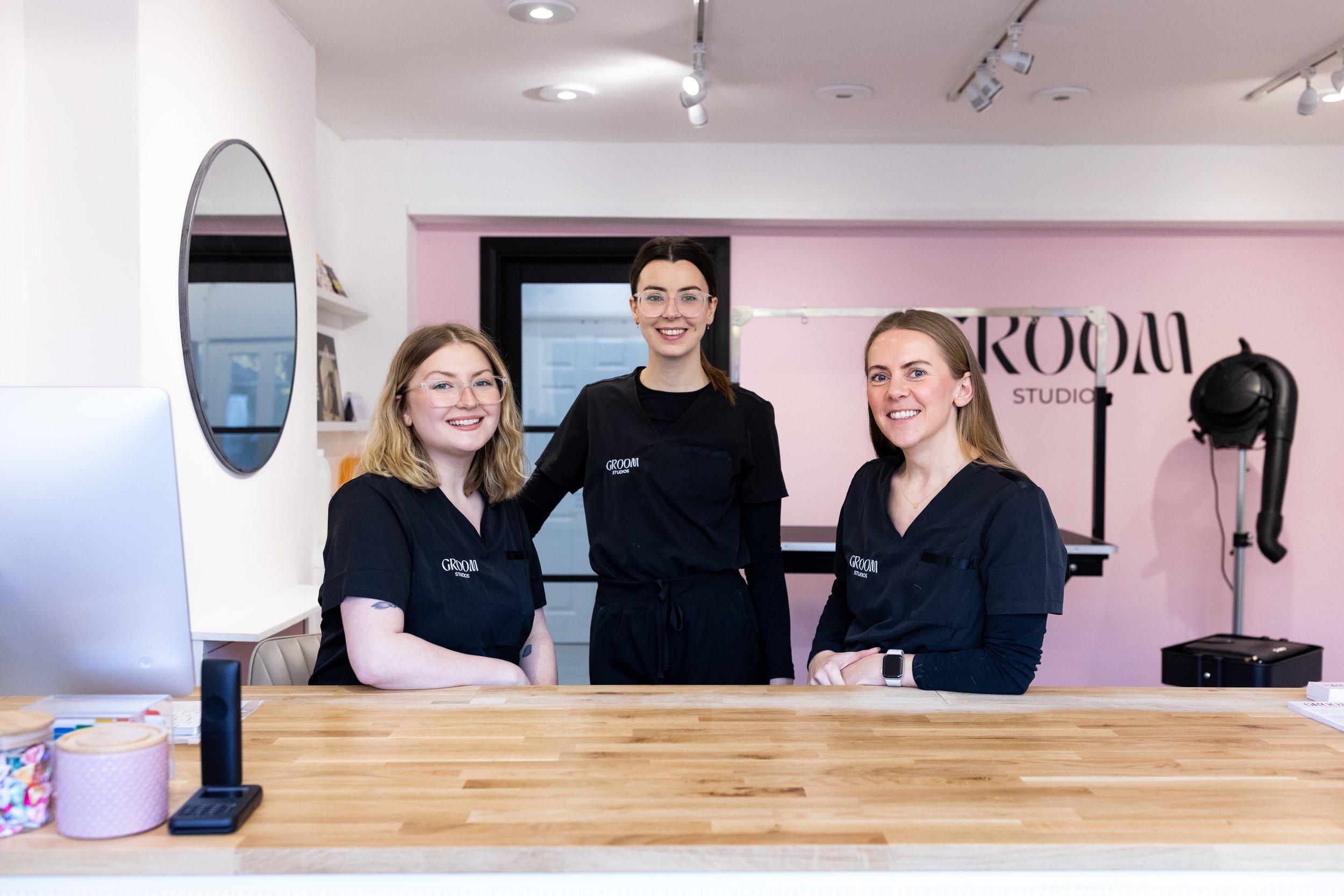 Three women in black uniforms standing at a reception desk smiling in a room with pink walls and passport equipment in the background.