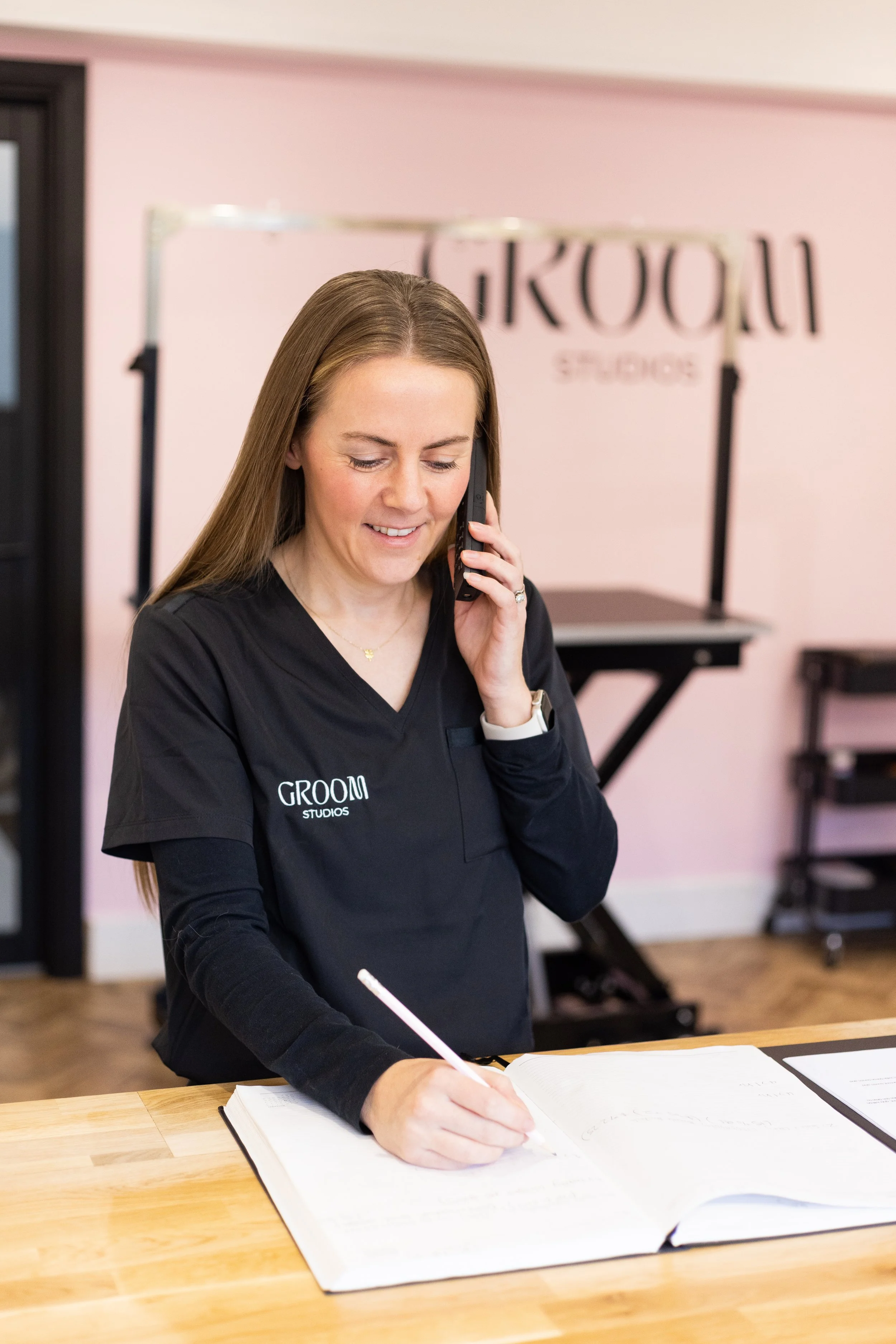 A woman in a black shirt with 'GROOM STUDIOS' written on it, smiling while talking on a cell phone and writing in a guest book at a studio reception area.