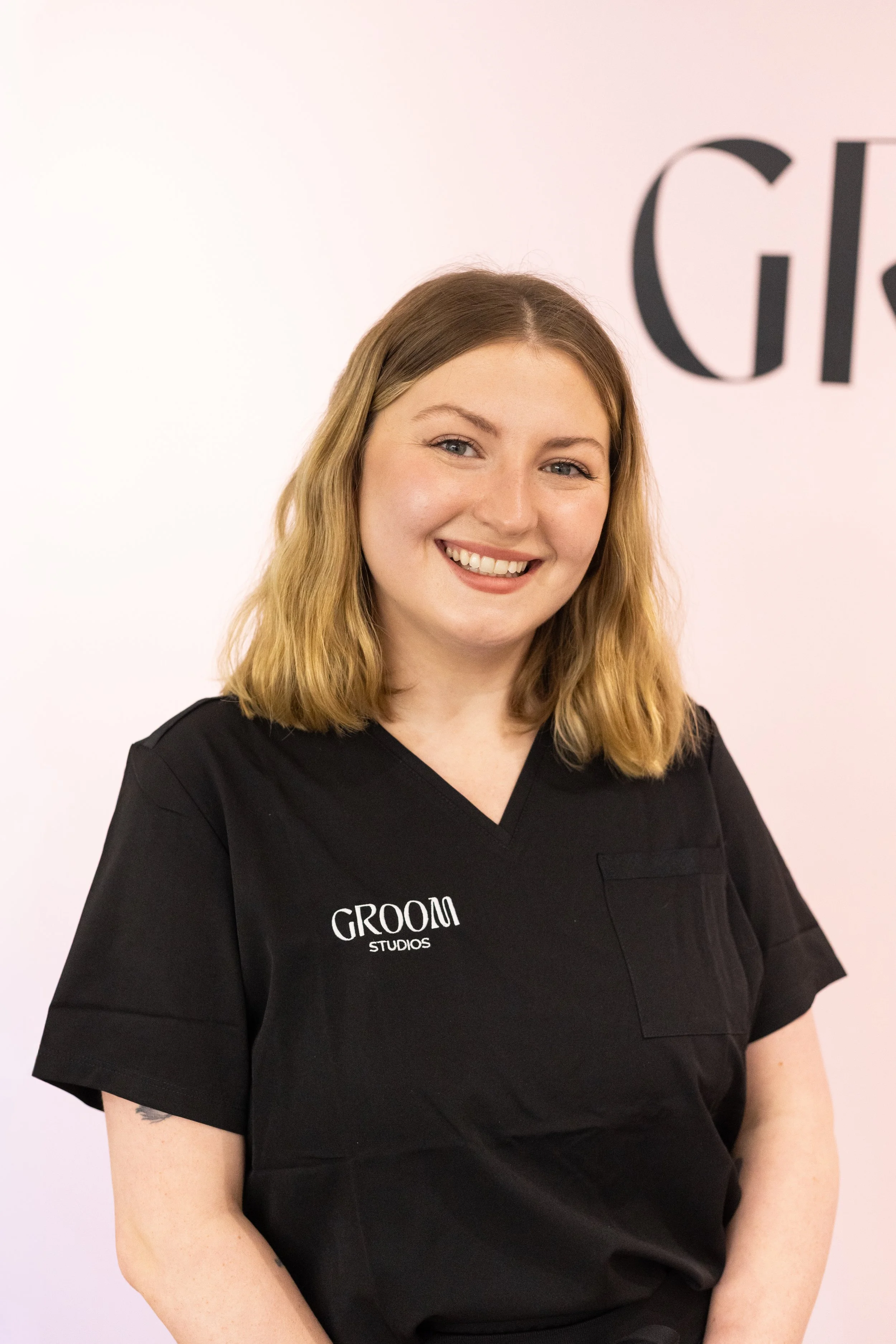 A woman with shoulder-length blond hair smiling, wearing a black scrubs top with 'GROOM STUDIOS' embroidered on it, standing in front of a light pink wall with partially visible black text.
