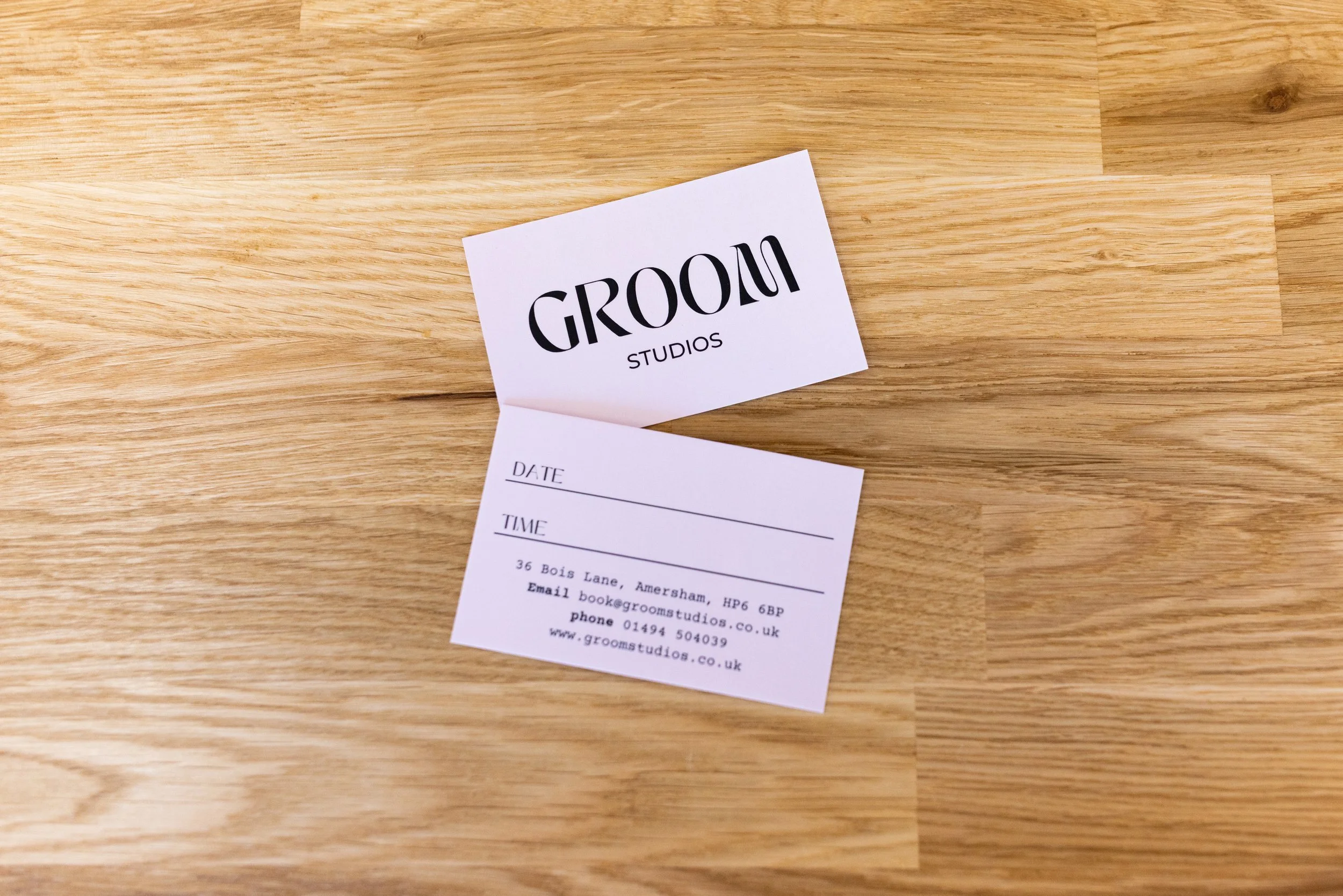 A printed business card for Groom Studios, placed on a wooden surface. The card includes spaces for date and time, and provides contact information including an address, email, phone number, and website.