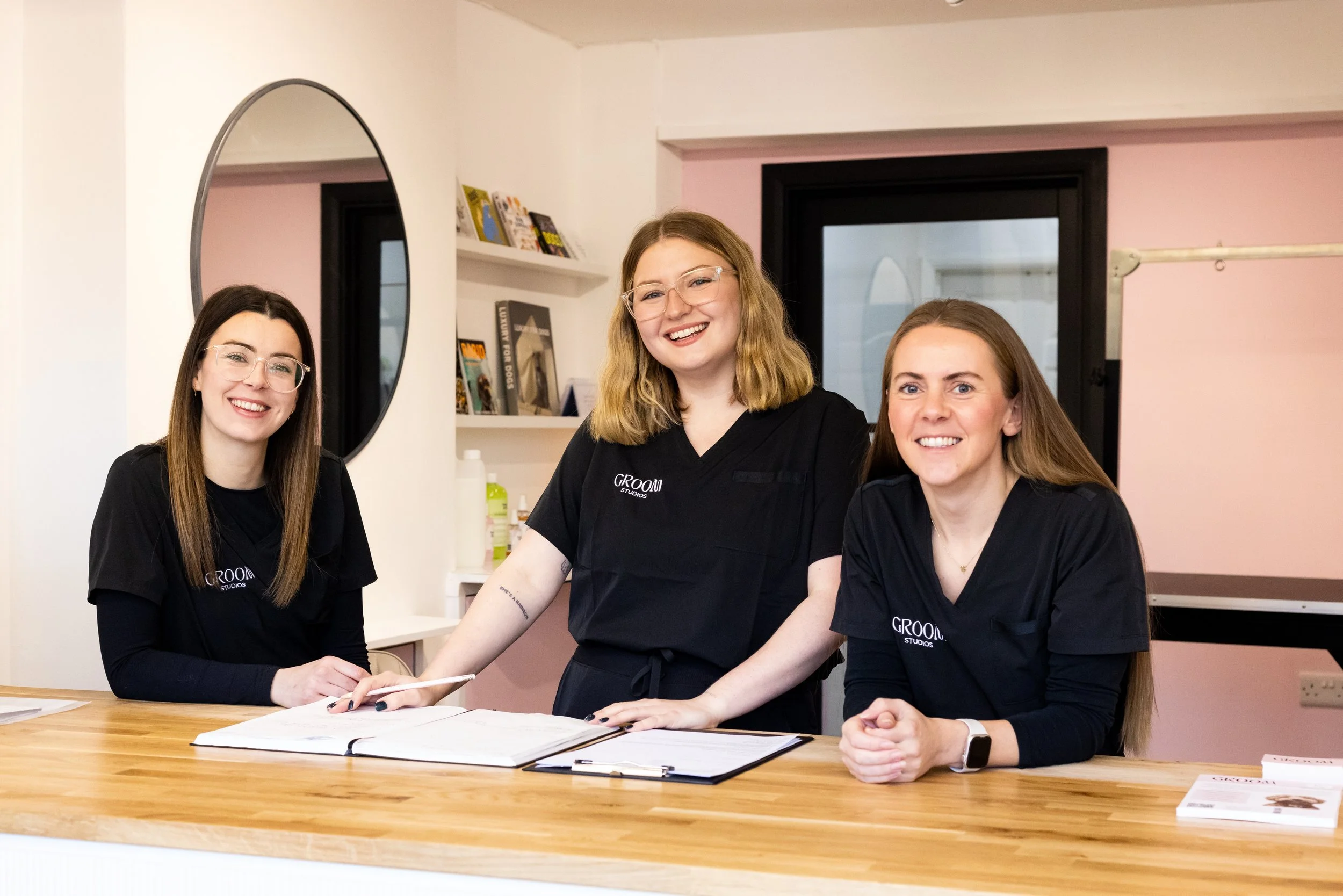 Three women in black T-shirts with 'GROOM STUDIOS' logo, smiling behind a wooden reception counter, with a mirror, shelves, and a pink wall in the background.